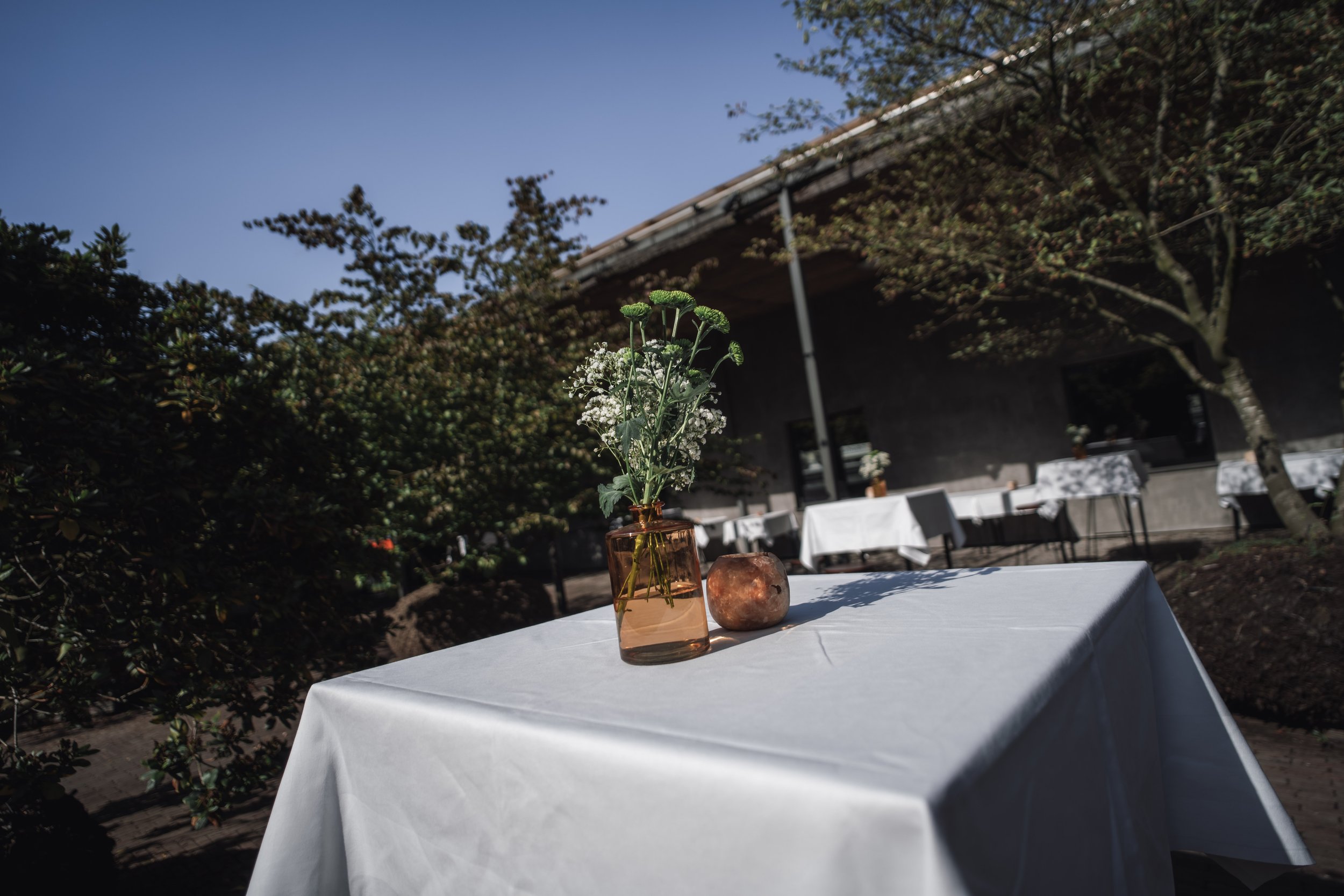 A table with a white tablecloth outdoors, holding a small vase with green and white flowers and a salt lamp, set in a garden area with trees and a house in the background under a clear blue sky.