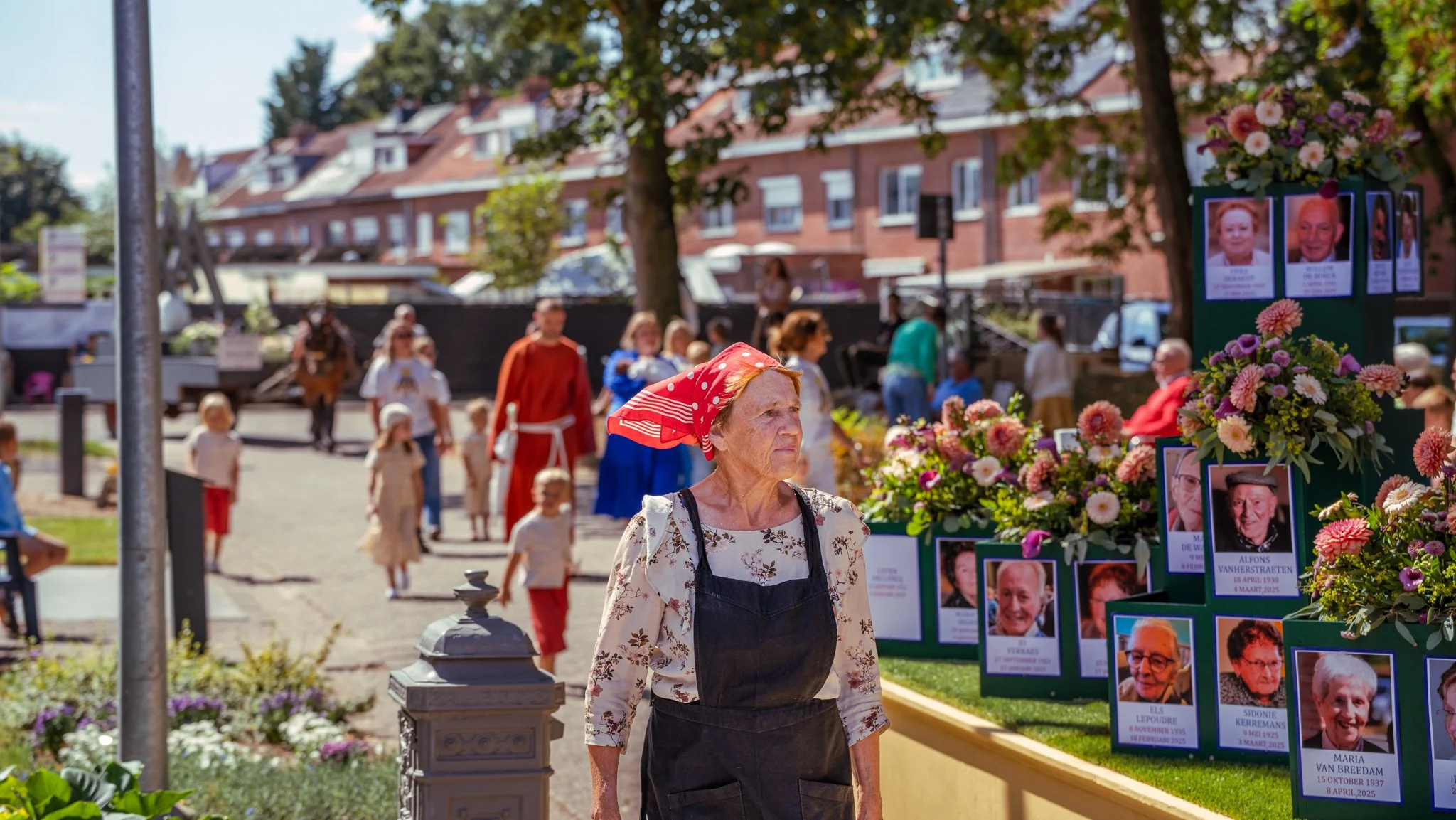 An elderly woman wearing a red headscarf and a black apron stands near a memorial display with photos and flowers, with a crowd and buildings in the background.