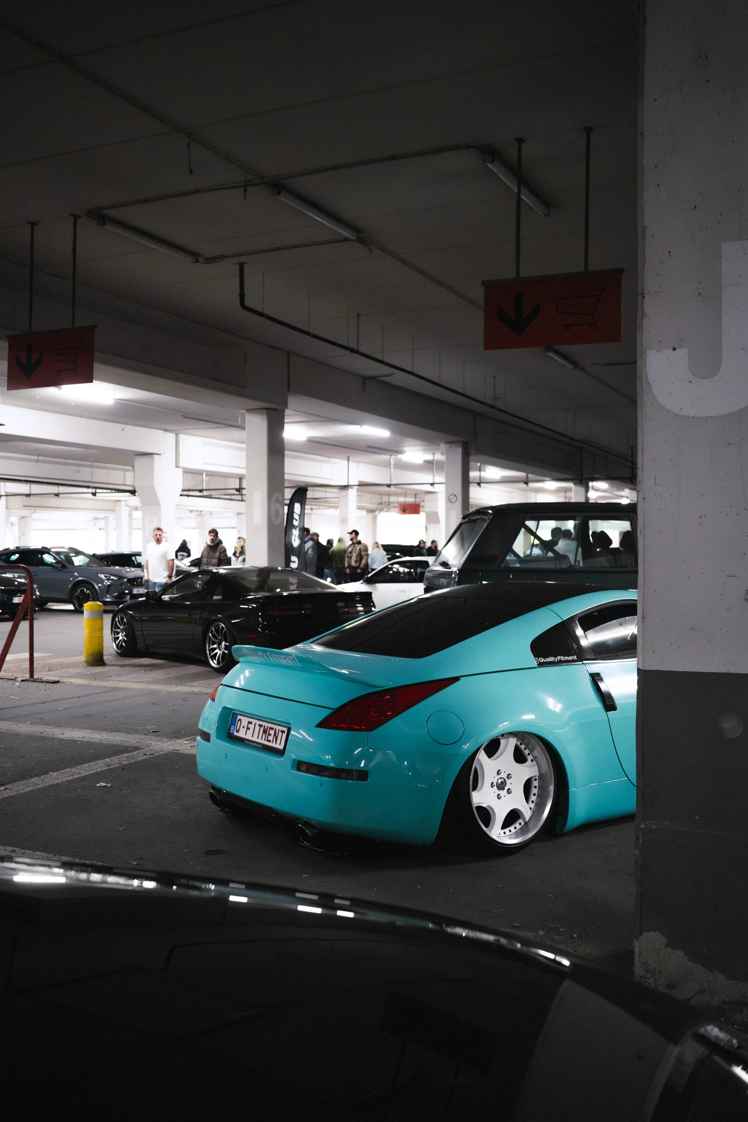 A parking garage filled with various cars, including a prominent light blue sports car with custom wheels and a license plate reading 'Q-FITMENT'. There are people walking around and signs hanging from the ceiling indicating directions.