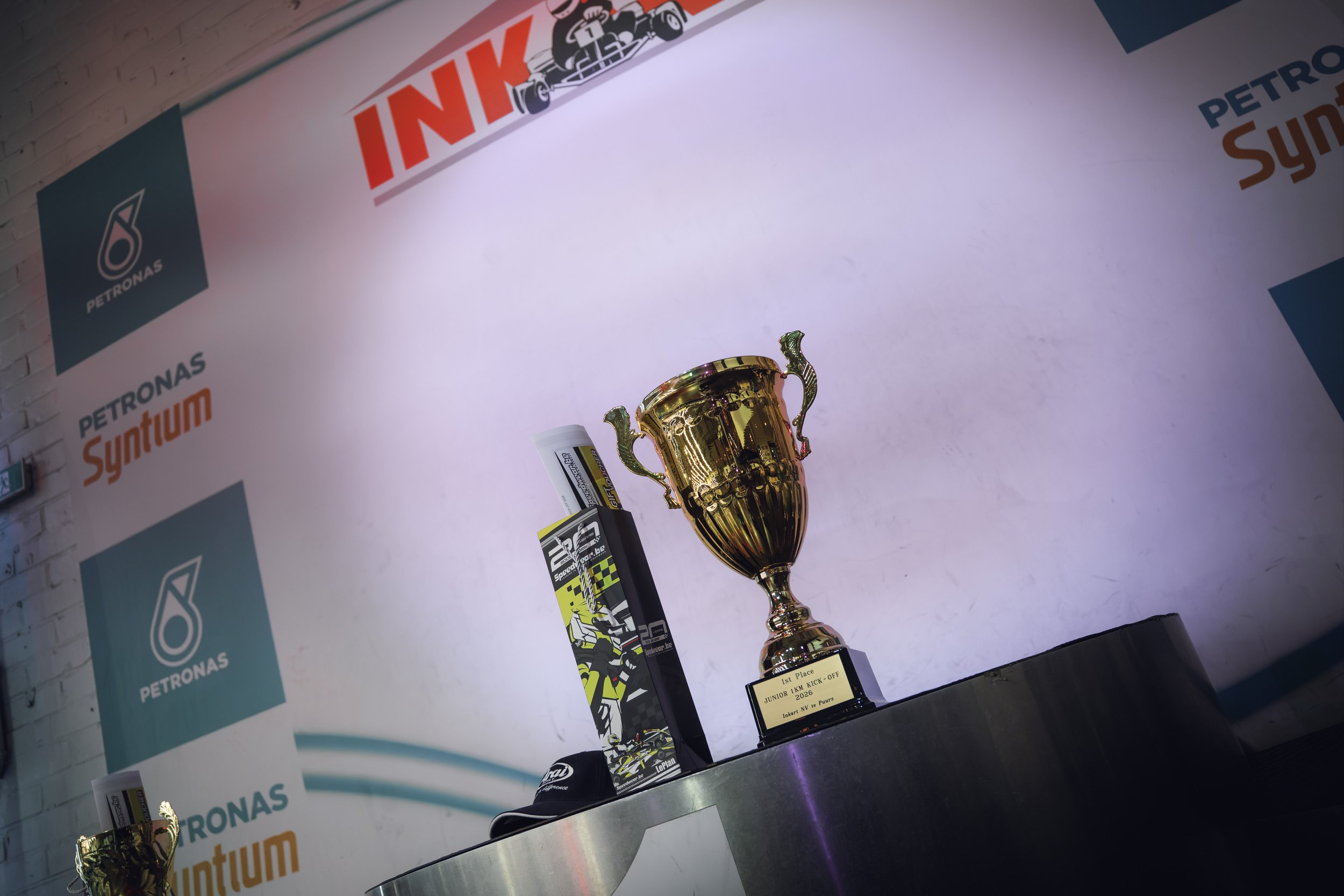 A gold trophy award on a table with a label indicating first place in a junior kick-off event in 2020. The background features logos of Petronas and other sponsors, and a poster with a racing theme.