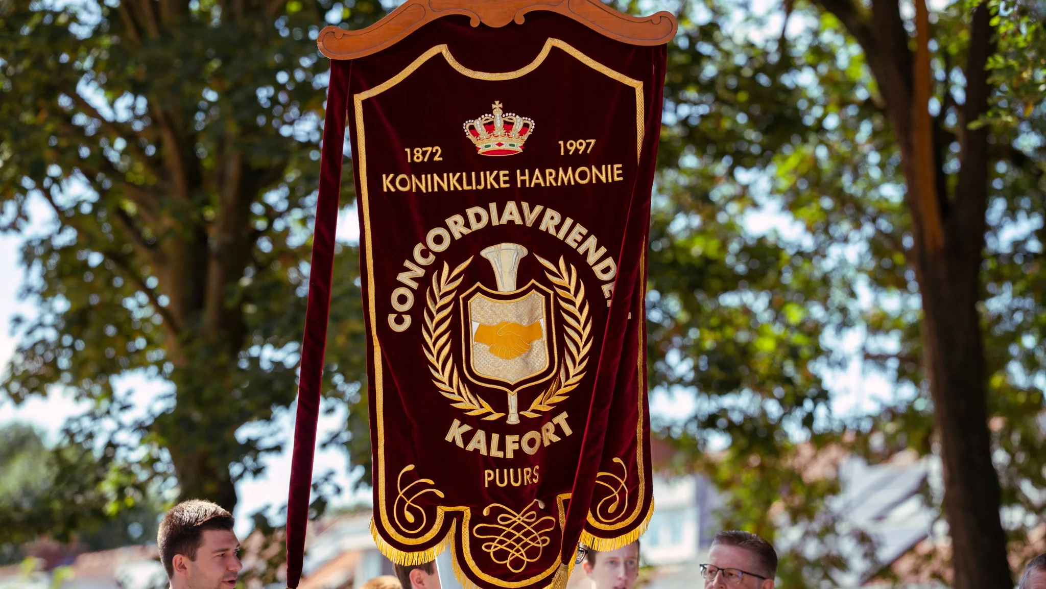 A ceremonial banner with a crown and handshake emblem, read 'Koninklijke Harmonie Congeniaanvriend Kalfort,' celebrating 125 years from 1872 to 1997, held outdoors among people and trees.
