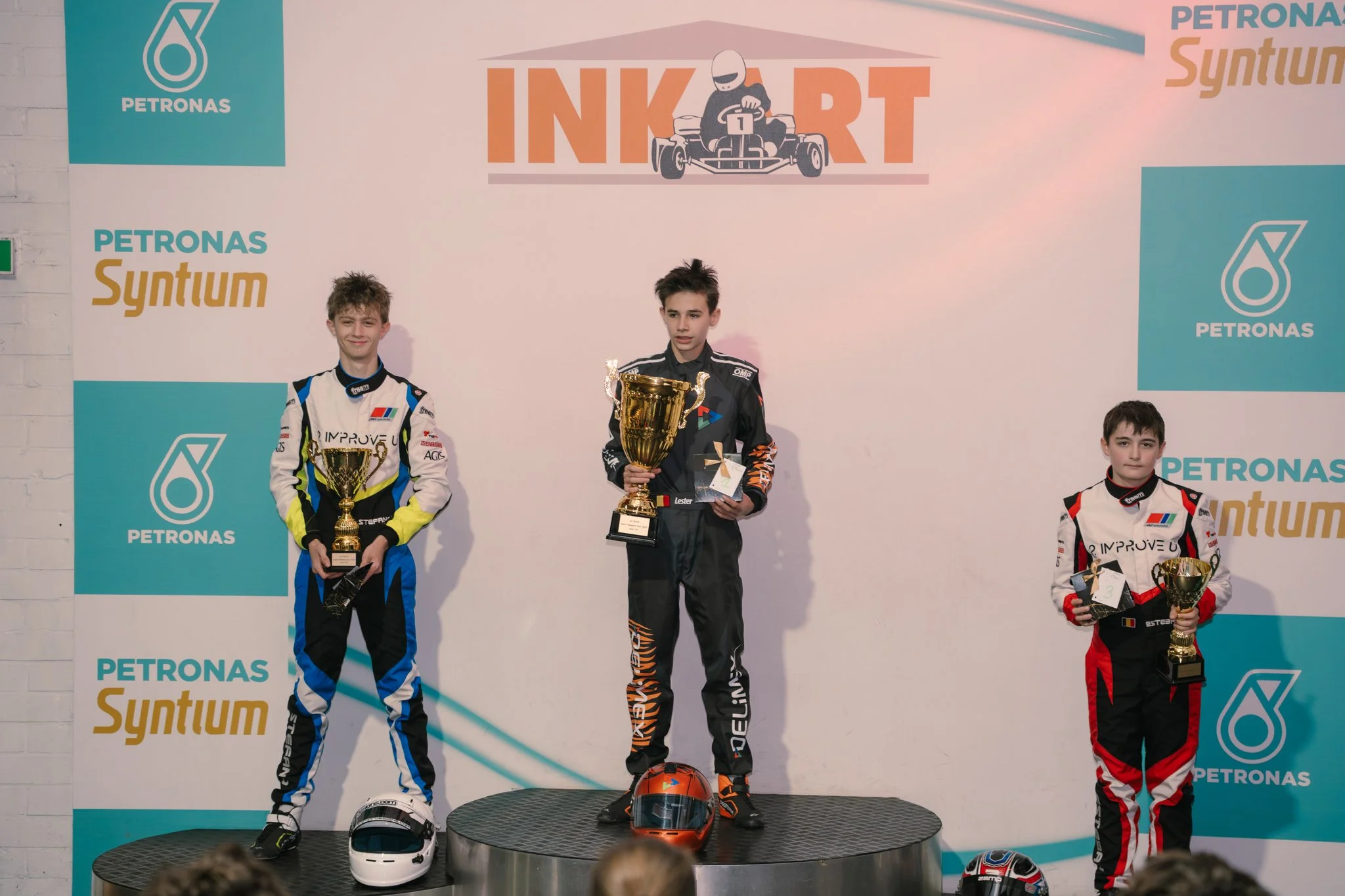 Three young race car drivers standing on winners' podium holding trophies, with the first-place winner in the center on the highest step, the second-place to his left, and the third-place to his right. All are in racing suits, and there are helmets o