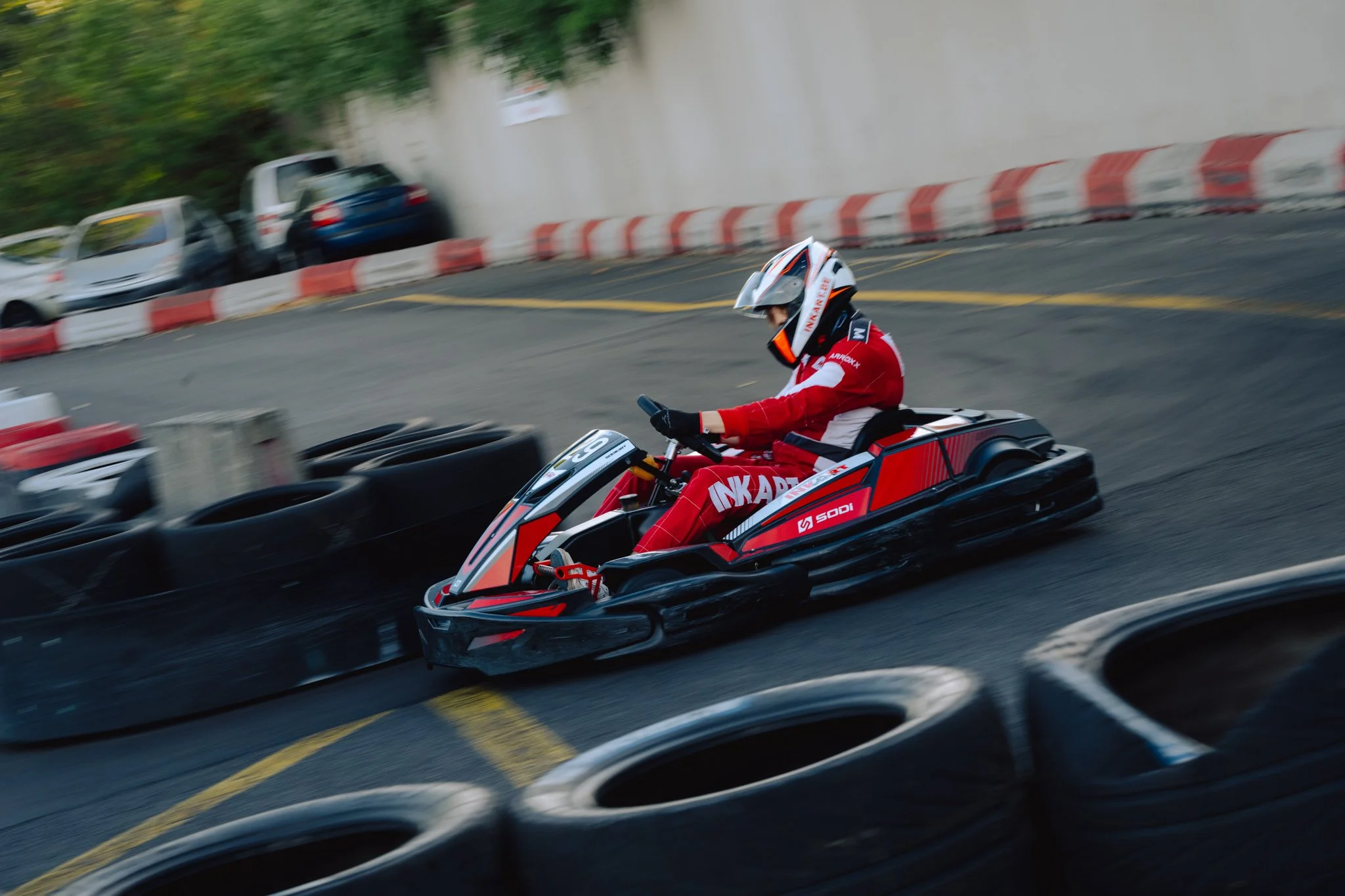 A person racing go-kart in a curve on an outdoor track, wearing a red racing suit and a helmet, with tires and cars parked nearby.