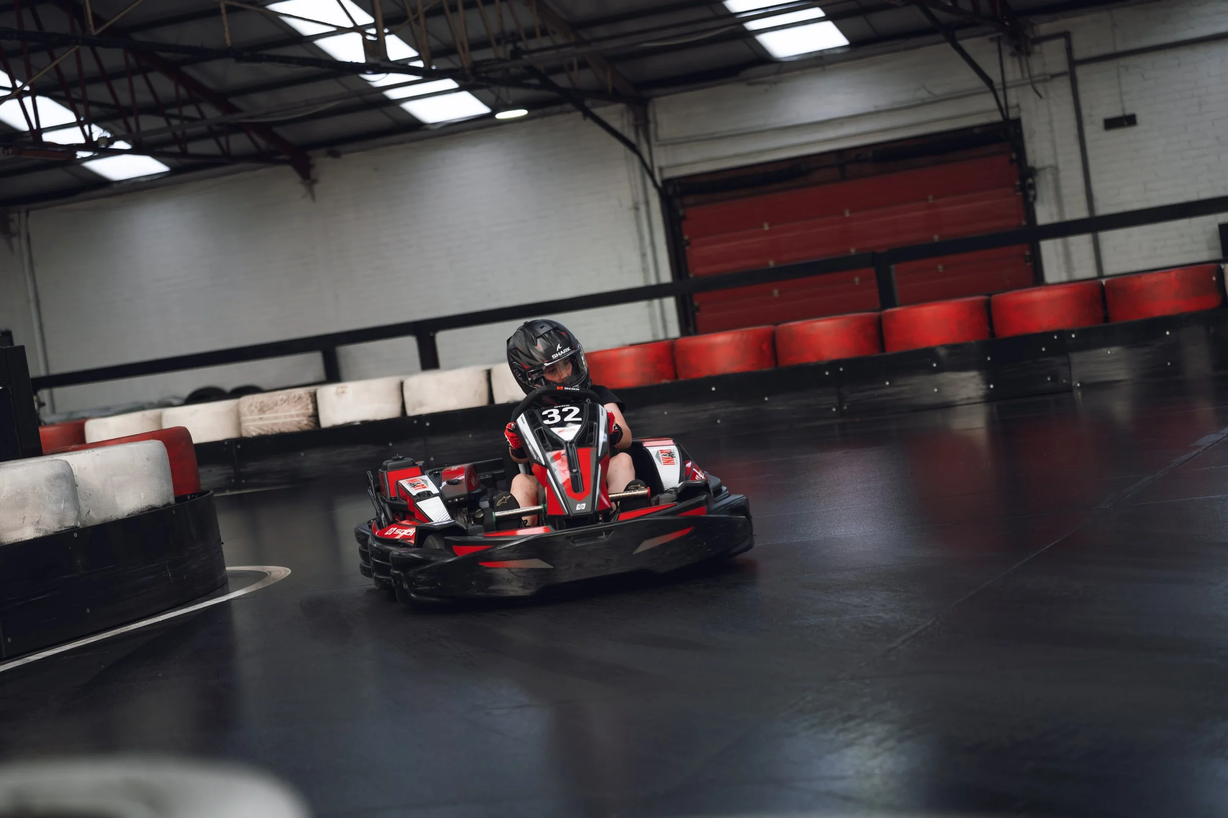 A young person wearing a helmet driving a red and black go-kart on an indoor racing track with barriers and a roof overhead.