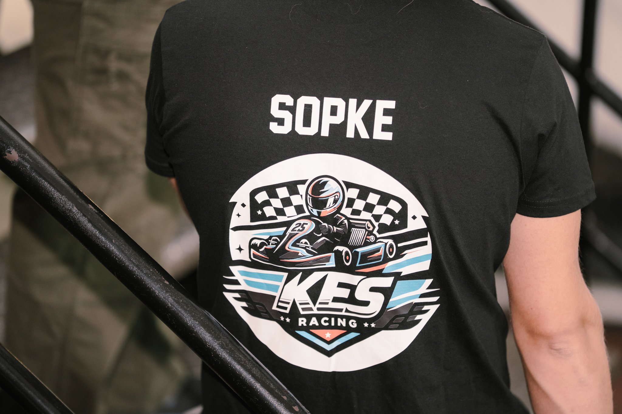 A person wearing a black T-shirt with a racing logo featuring a go-kart driver, checkered flag, and the words 'SOPKE KGS Racing'.
