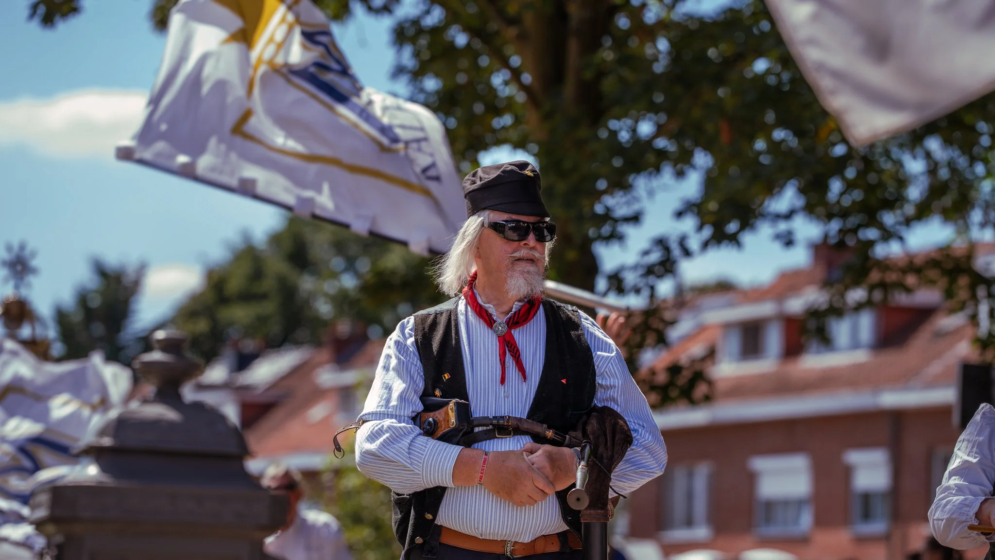 An elderly man with long gray hair and beard, wearing sunglasses, a black hat, a red neckerchief, and a striped shirt, holding a walking stick, standing outdoors during daytime with umbrellas and trees in the background.