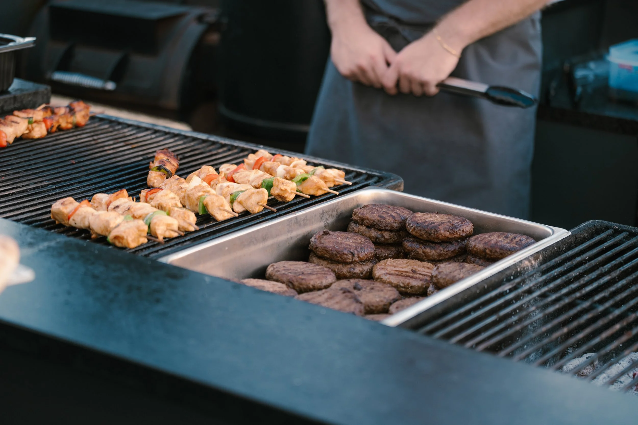 Close-up of grilled skewers with chicken and vegetables, and hamburger patties on a barbecue grill, with a person's hands holding tongs in the background.