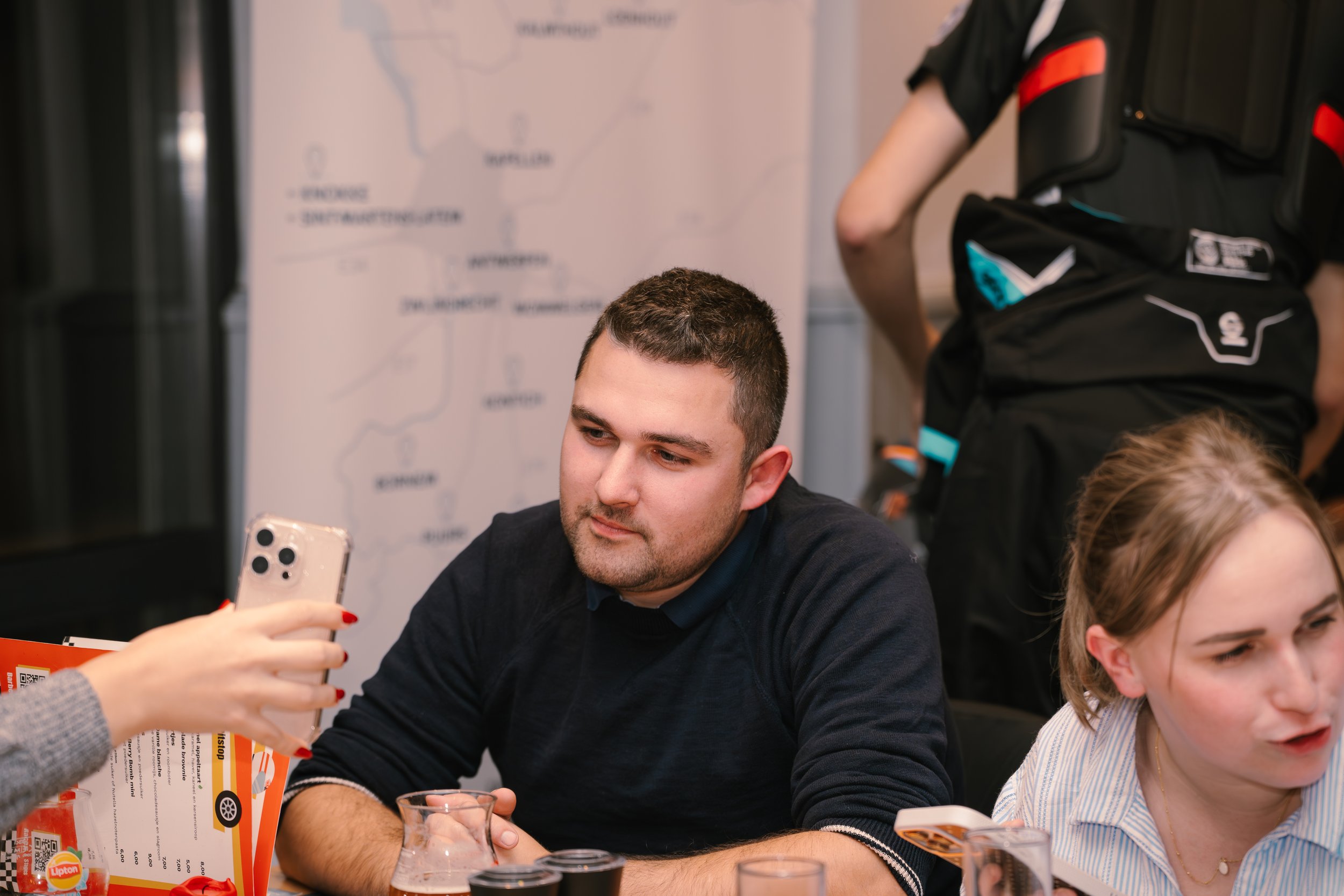 A young man with short dark hair and a beard looking at his phone while sitting at a table with others. A woman in a gray sweater is handing him a menu or flyer. In the background, there are posters and a person in a black uniform with red and blue a