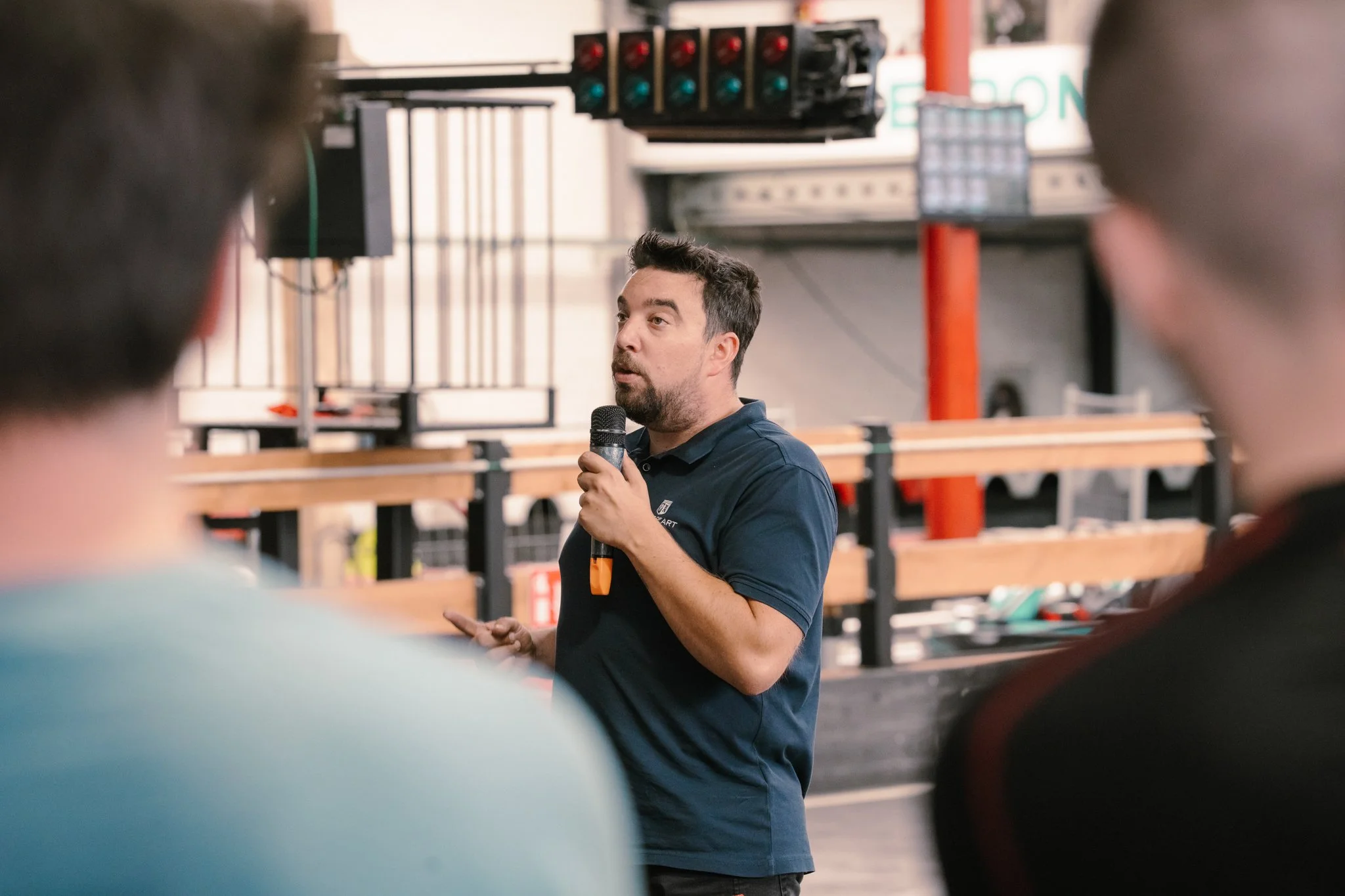A man speaking into a microphone during a presentation, with two people seen from behind in the foreground, in an industrial or workshop setting.