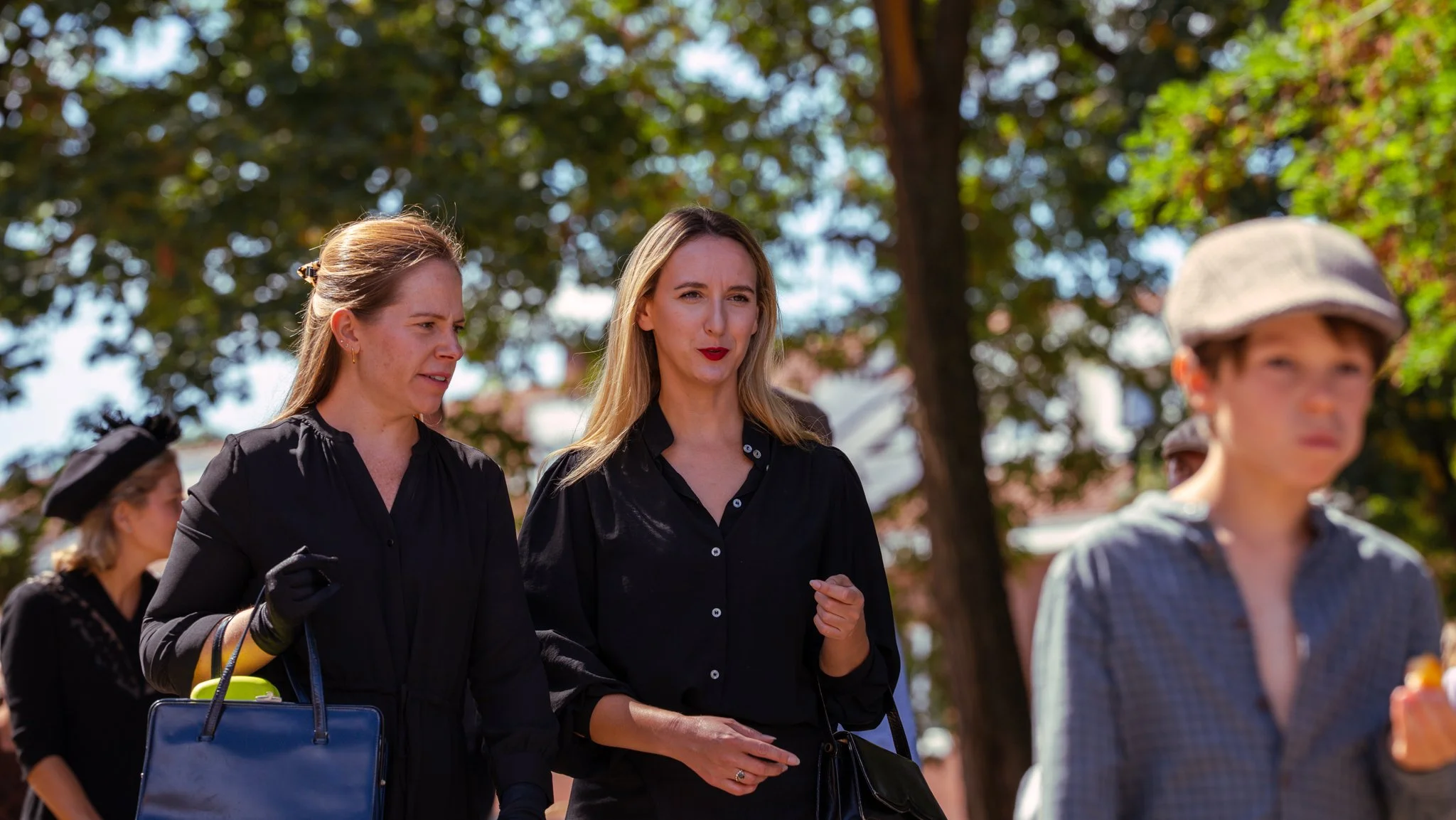 Group of women and a young boy walking outdoors with trees in the background. The women are dressed in black and one woman has blonde hair and red lipstick.