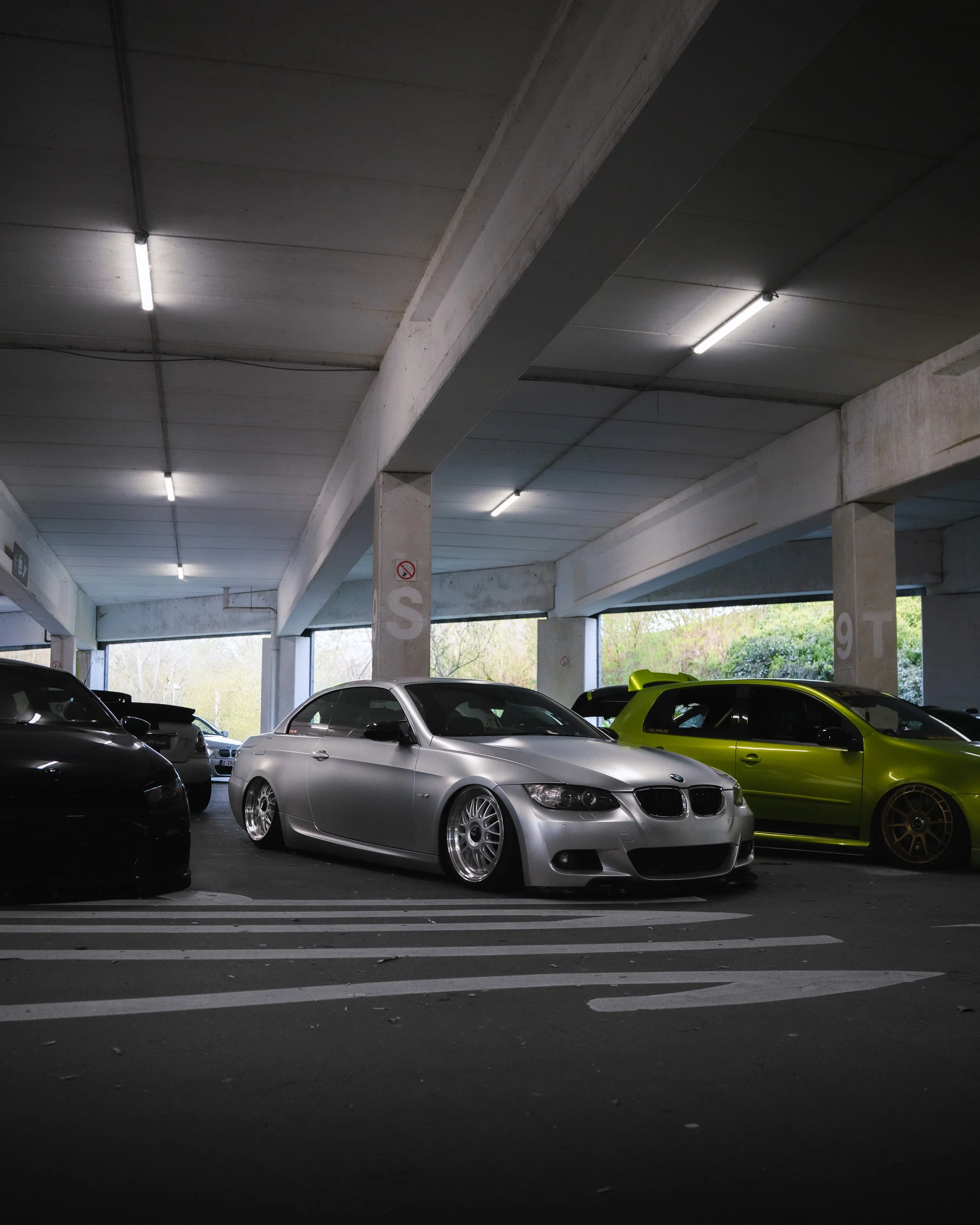 Inside a parking garage with a concrete ceiling and multiple cars parked, including a silver BMW, a green car, and others.