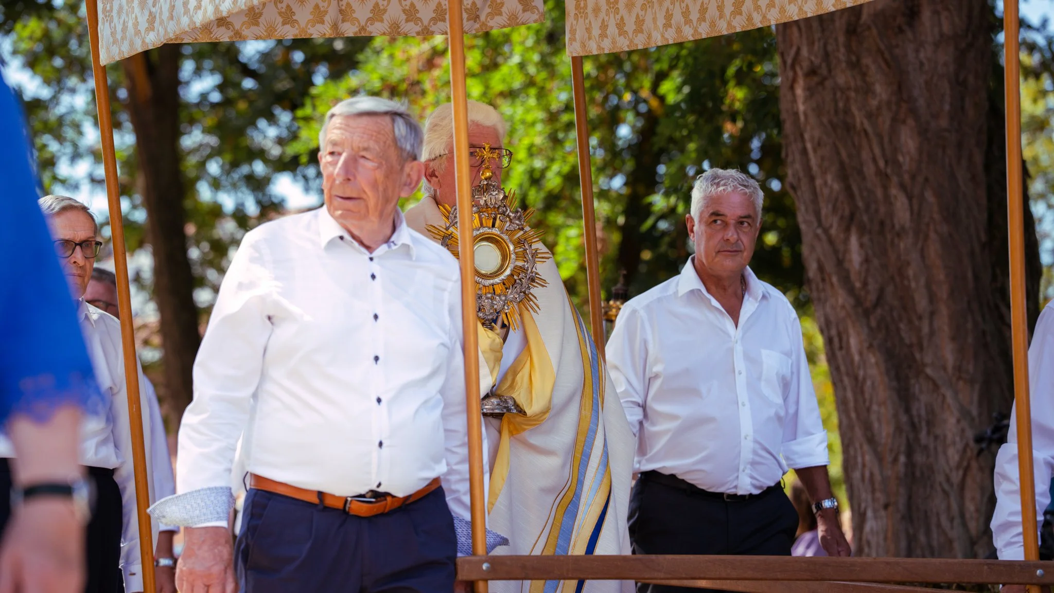 Group of men standing outdoors, one dressed in religious vestments, participating in a religious or cultural ceremony, with trees and a wooden structure in the background.