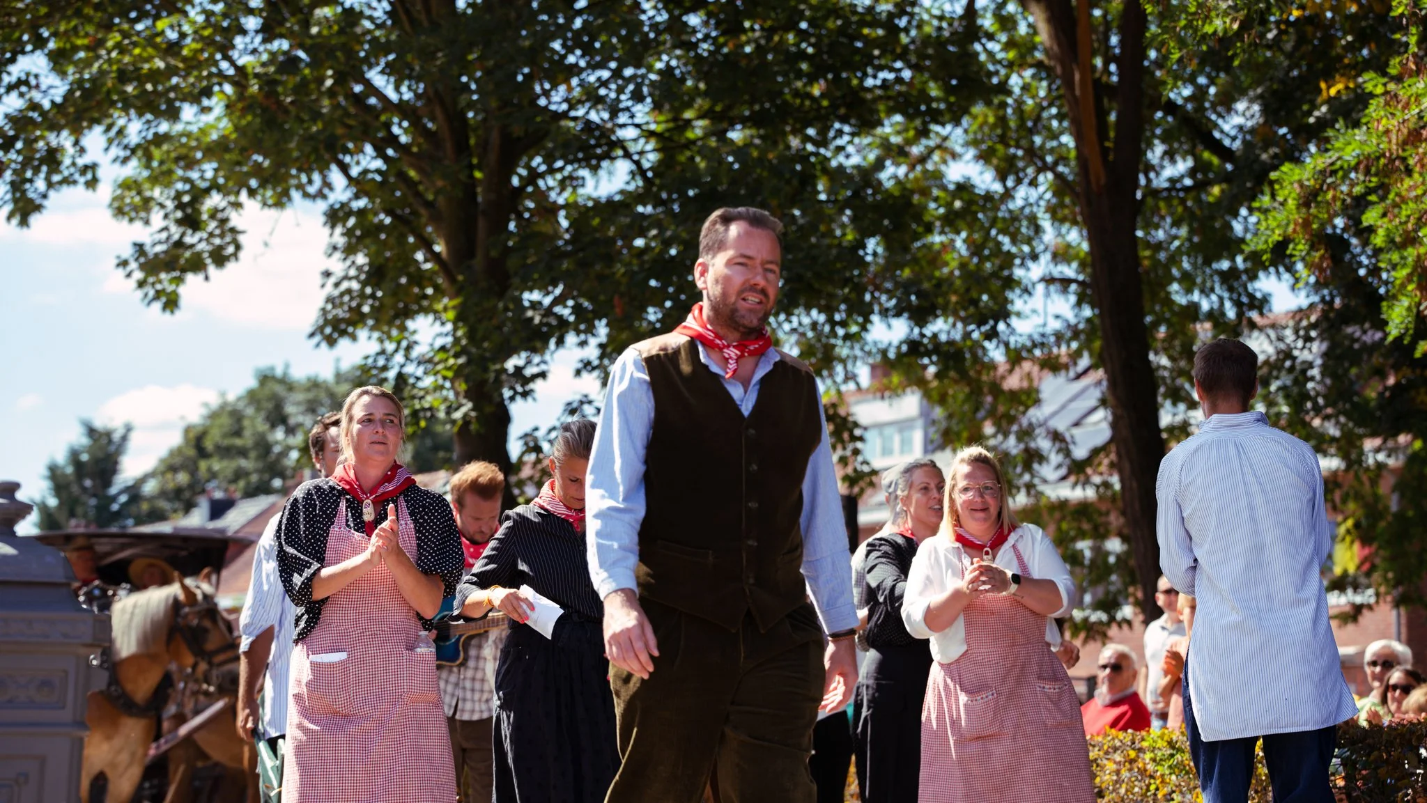 Group of people participating in a traditional outdoor event or celebration, wearing vintage or traditional clothing, with a focus on a man with a beard and a red scarf, standing under trees with a sunny sky in the background.