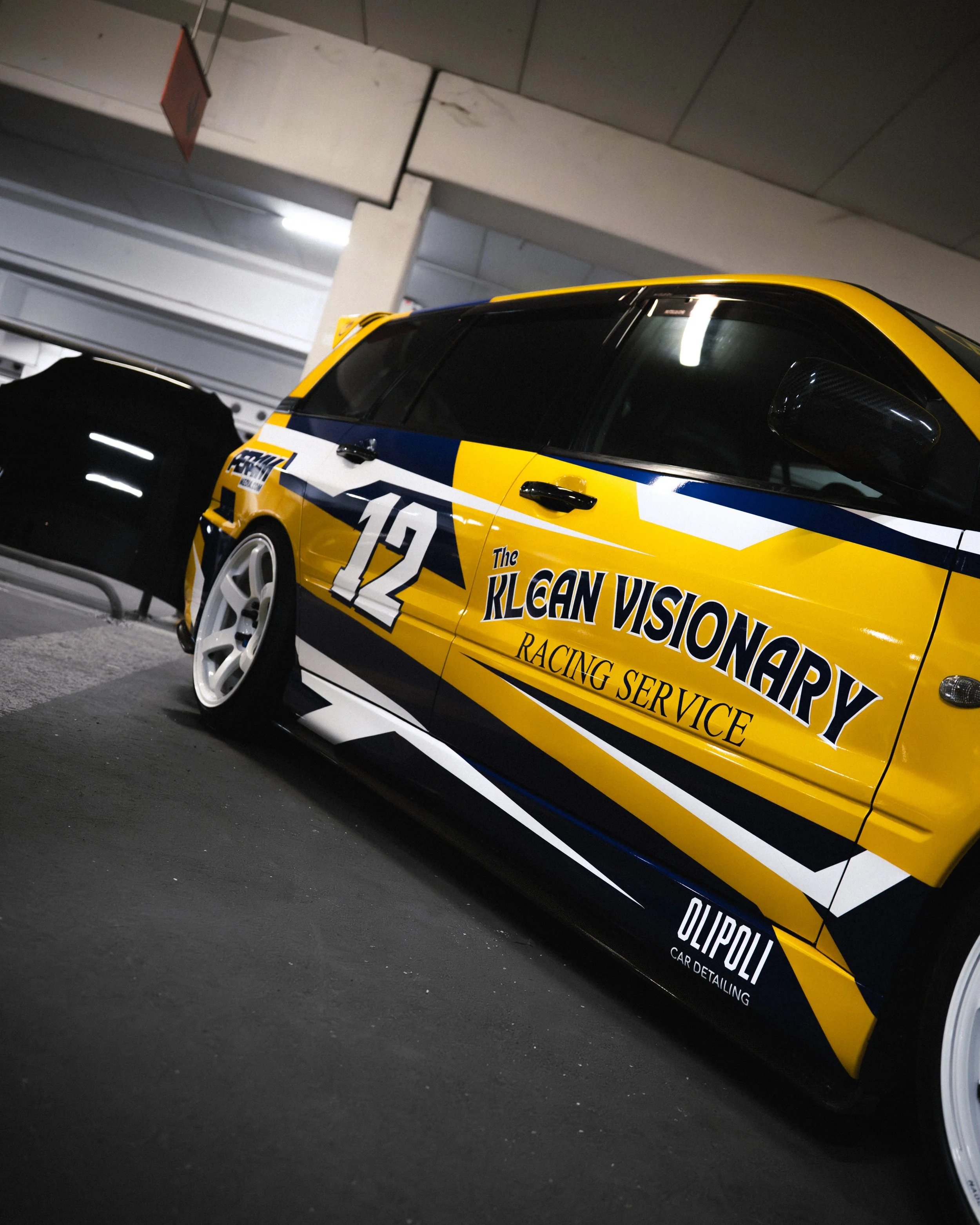 A yellow and black race car with sponsor decals, parked indoors in a parking garage, with a black folding chair beside it.