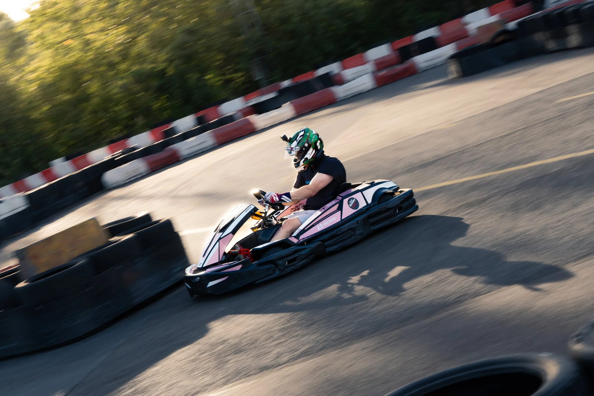 Person wearing a helmet with a GoPro camera mounted, riding a pink and black go-kart on an outdoor race track, with black and red tire barriers along the side.