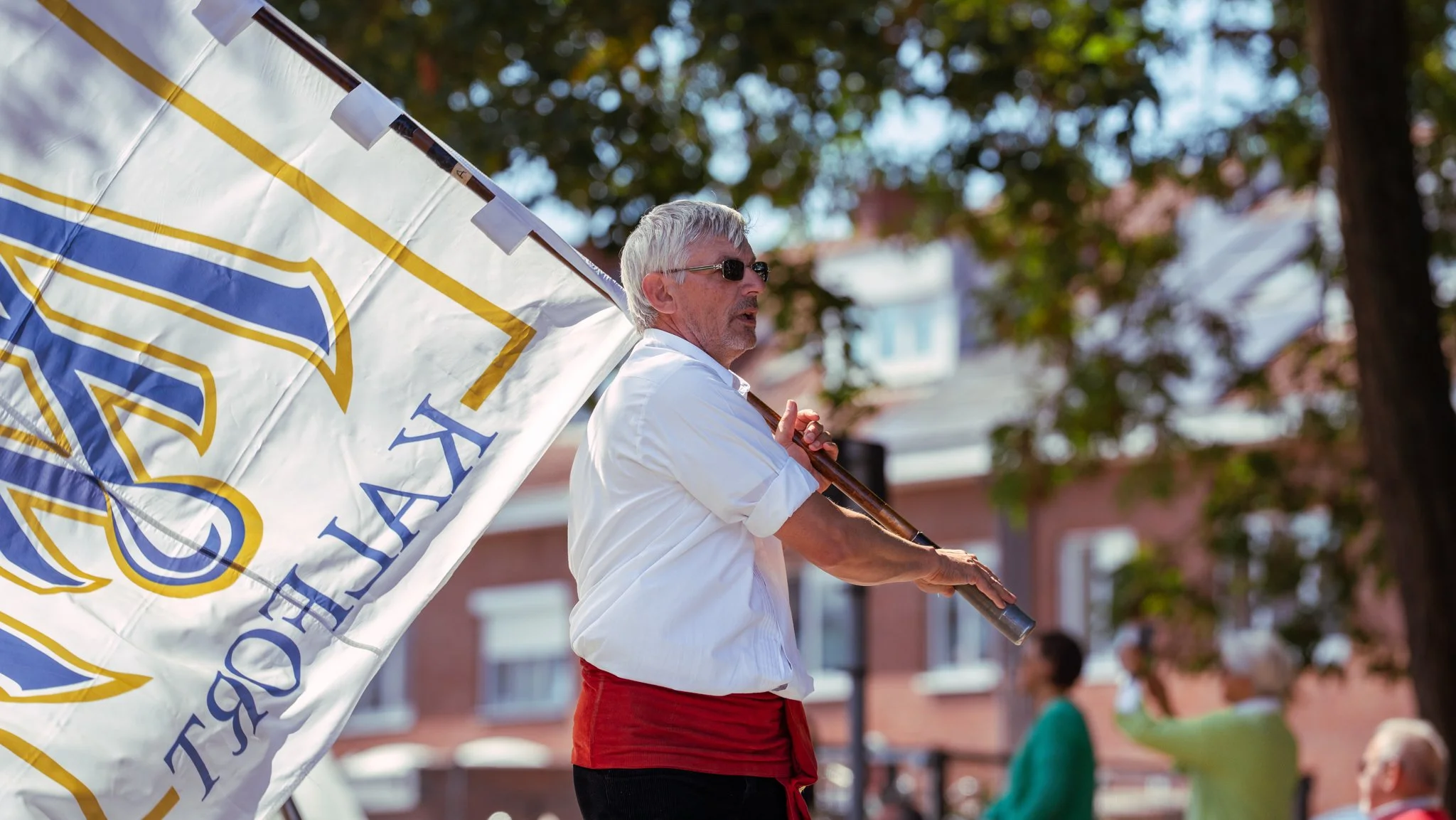 A middle-aged man with grey hair and glasses holding a large flag with the text 'JESUS' on it during an outdoor event, with trees and a brick building in the background.