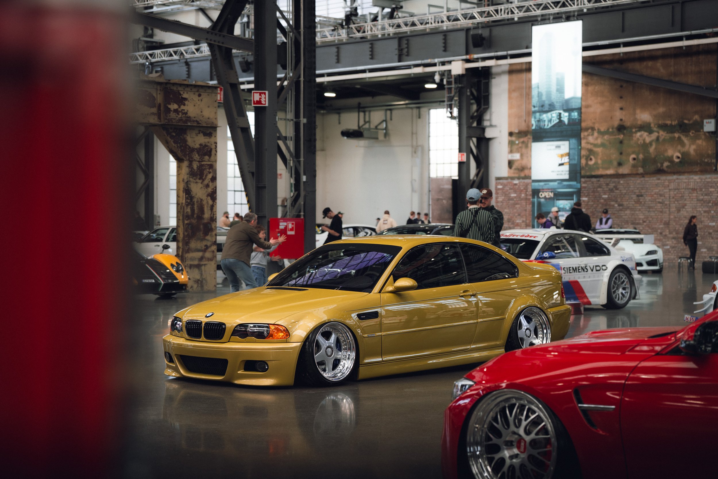 Indoor car exhibition showcasing a yellow BMW Z3 in the center, surrounded by red and white cars, with people observing and taking pictures, in a spacious industrial-style building with exposed brick and large windows.