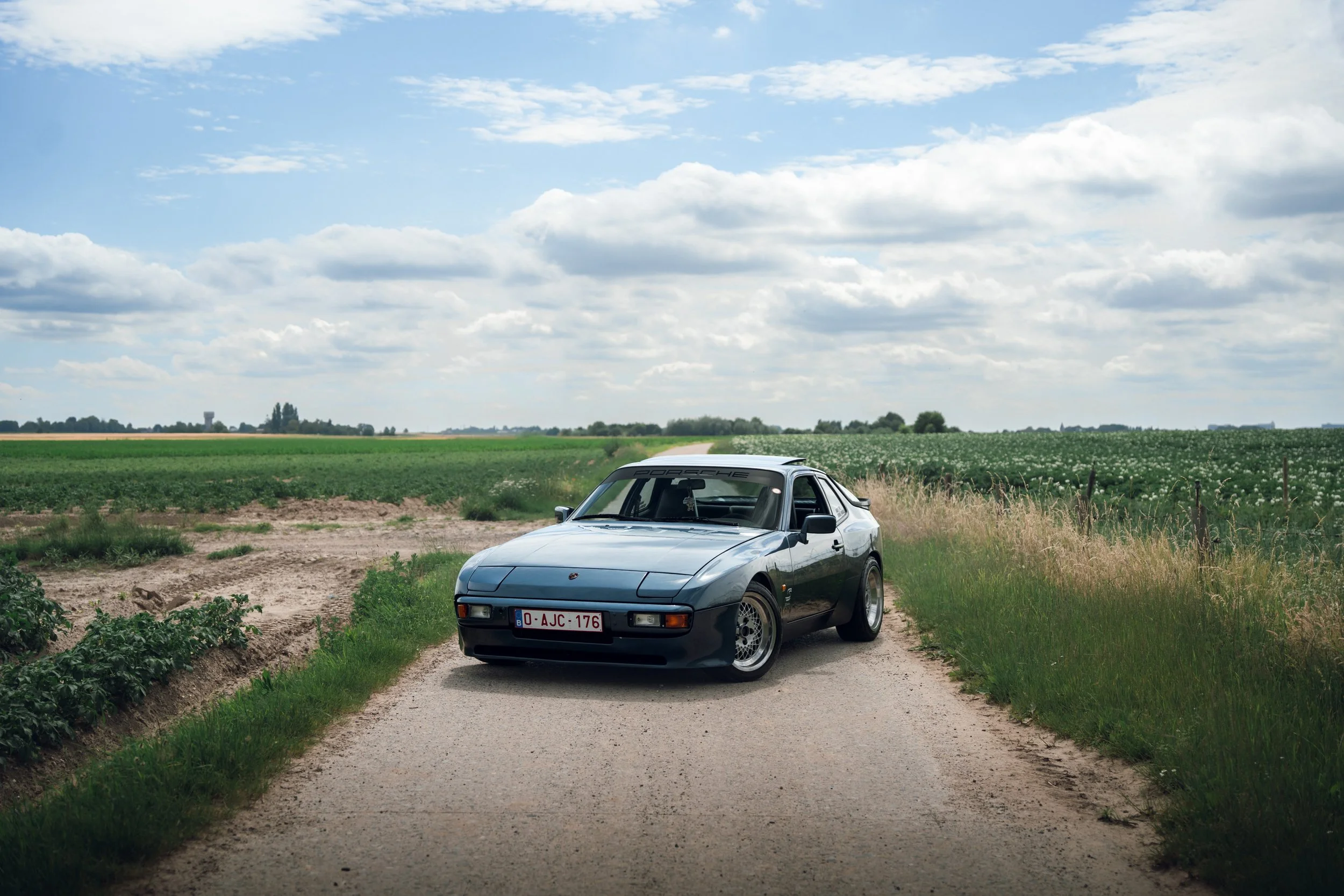 A vintage sports car parked on a narrow dirt road in a rural area surrounded by fields and greenery, with a cloudy sky overhead.
