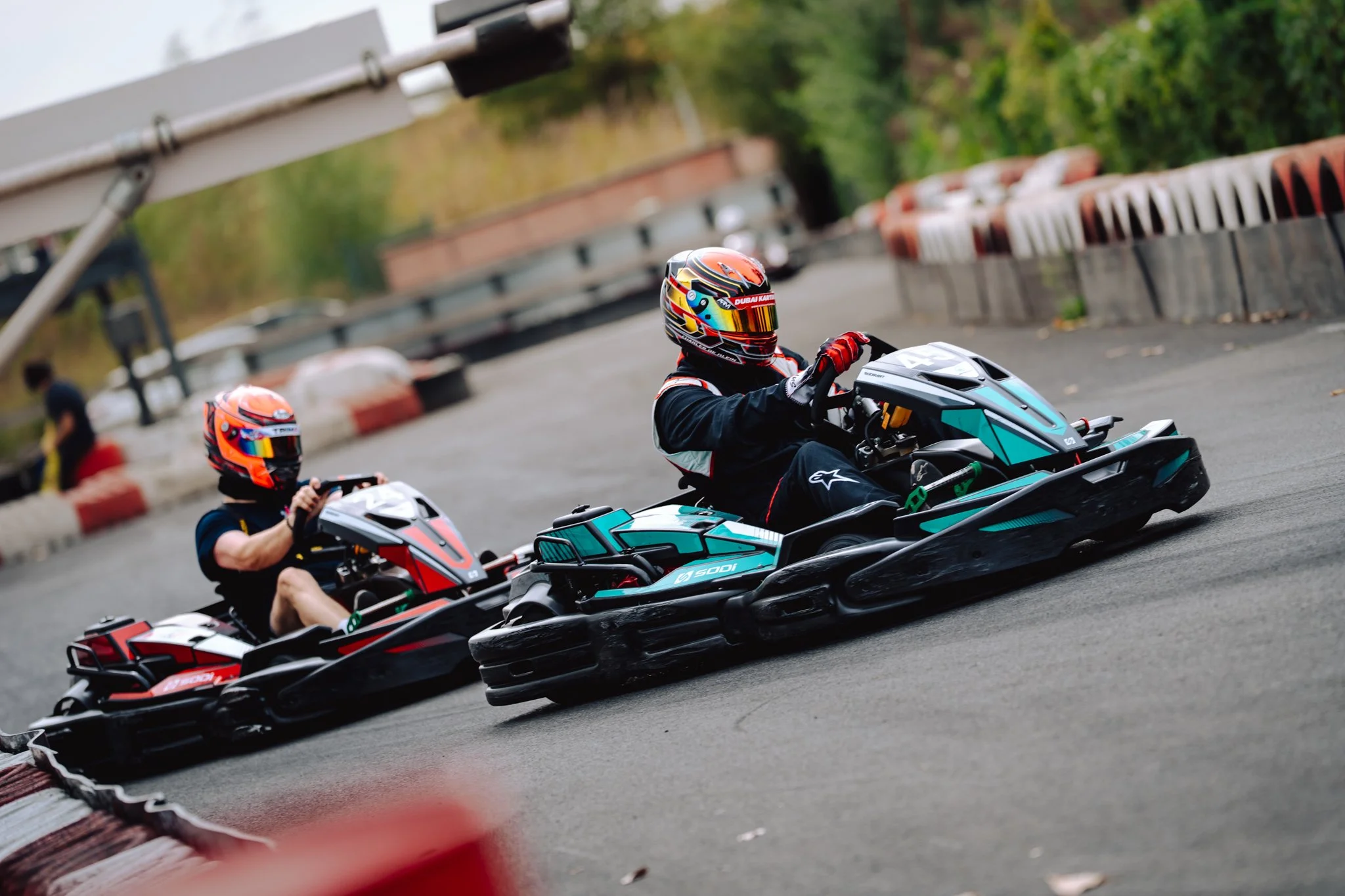 Two go-kart racers in black suits and colorful helmets, racing on an outdoor track with barriers and greenery.