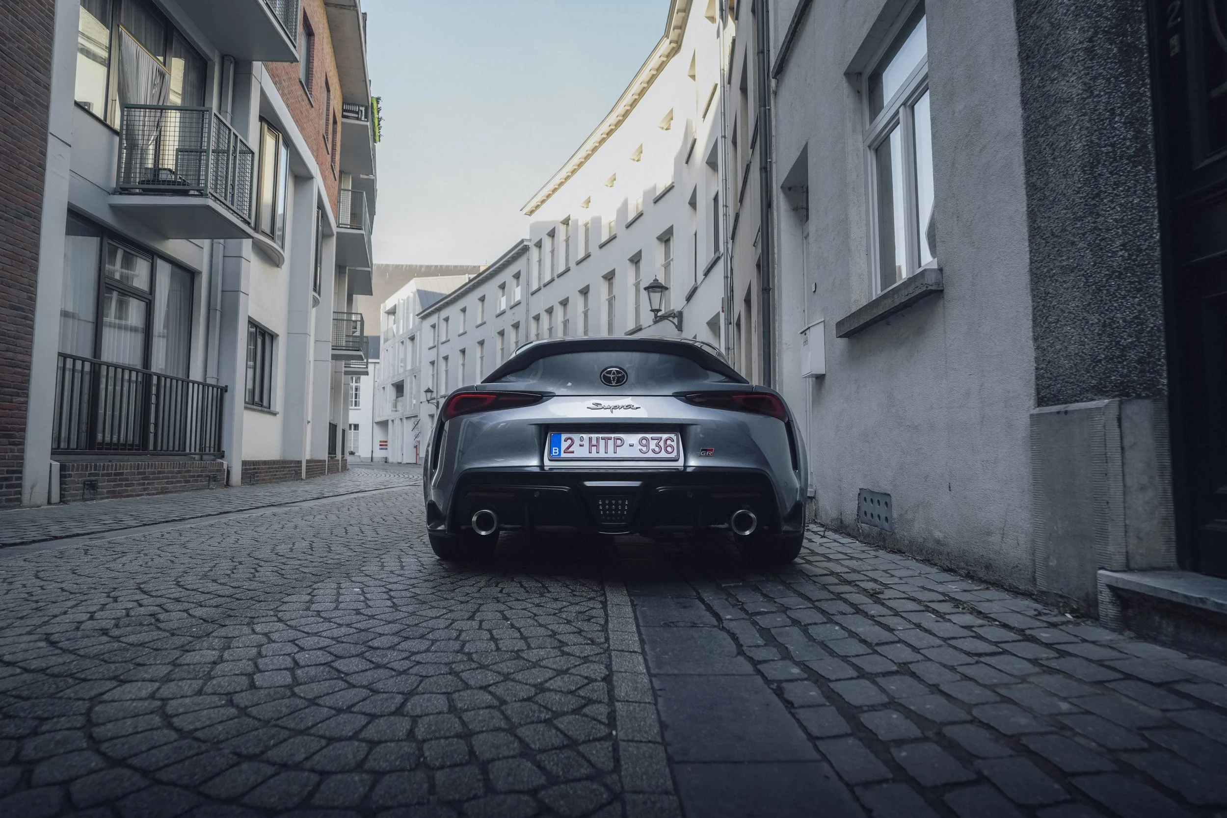 A gray Toyota Supra GR parked on a narrow cobblestone alleyway between white and gray residential buildings.