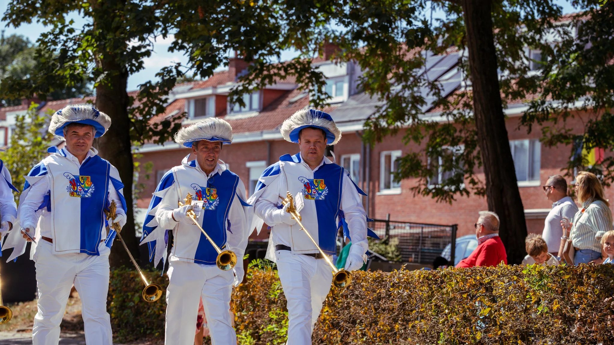 Four men dressed in white and blue costumes with hats, carrying brass horns, walking in a parade or festival on a sunny day, with spectators and trees in the background.