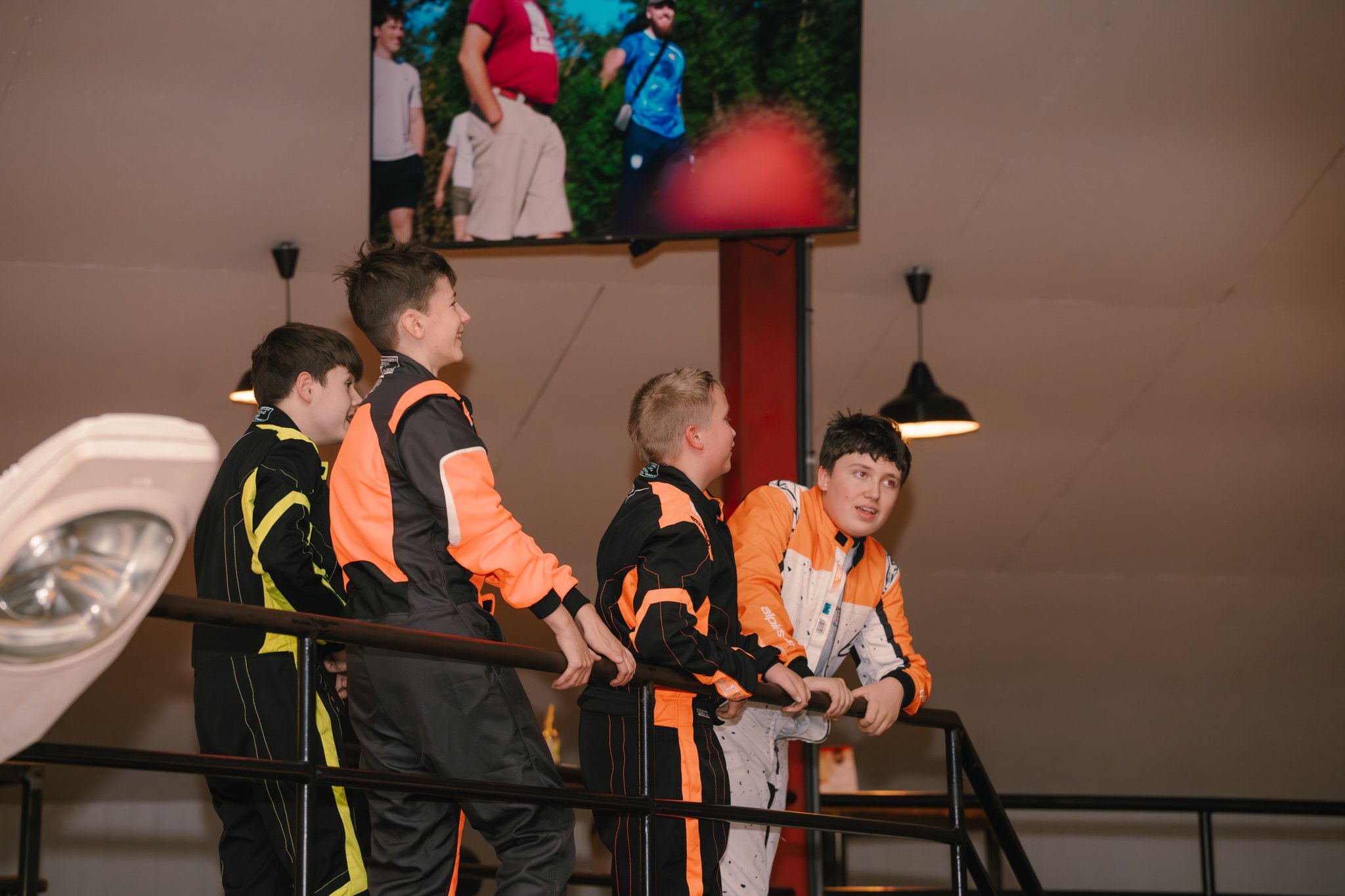 Four boys in racing suits stand behind a railing, watching a large screen showing a race or event with people outdoors.