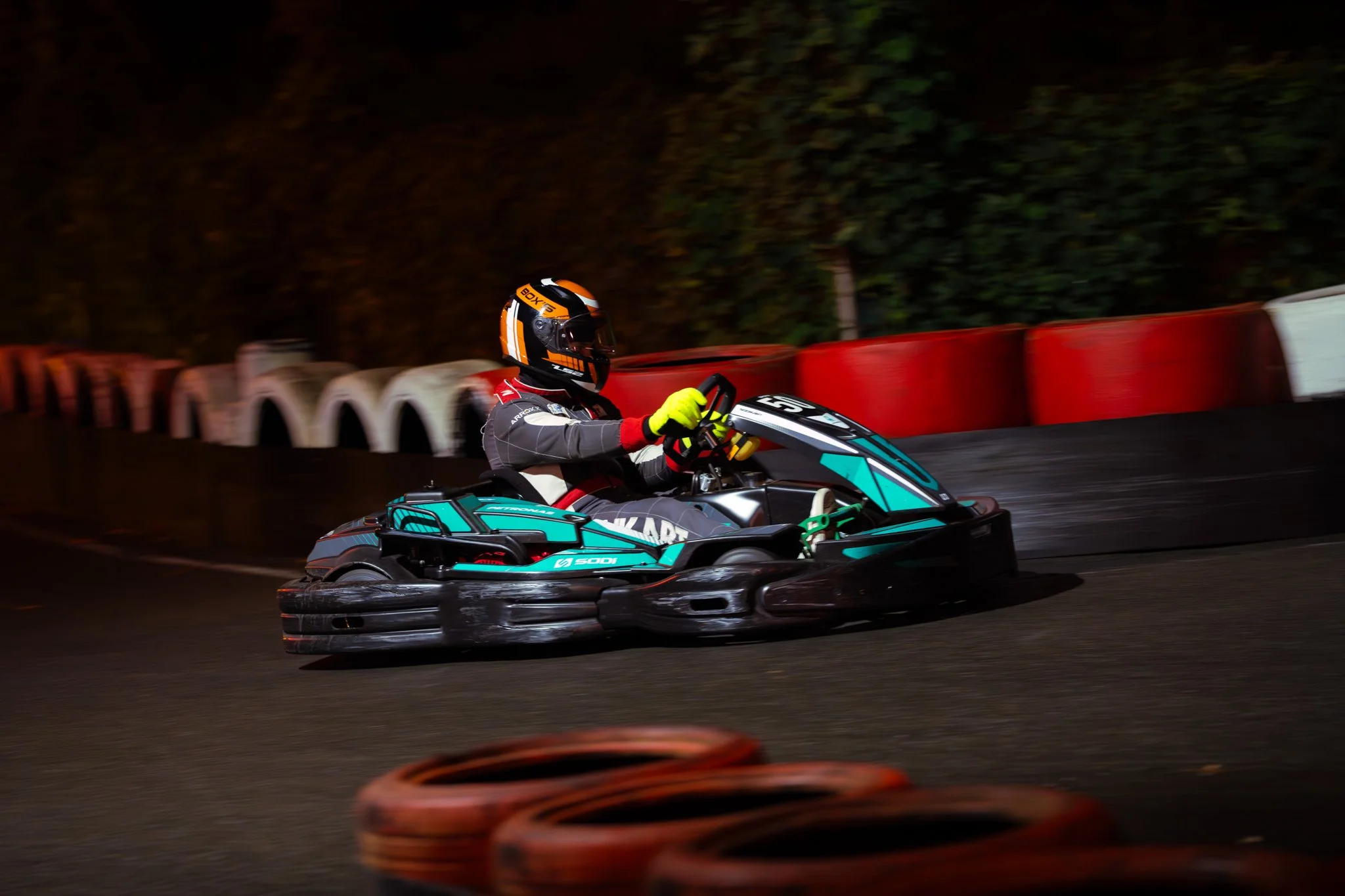 A go-kart racer wearing a black helmet, gray racing suit with red and white details, and bright yellow gloves, speeding along an indoor track with red and white tire barriers in the background.