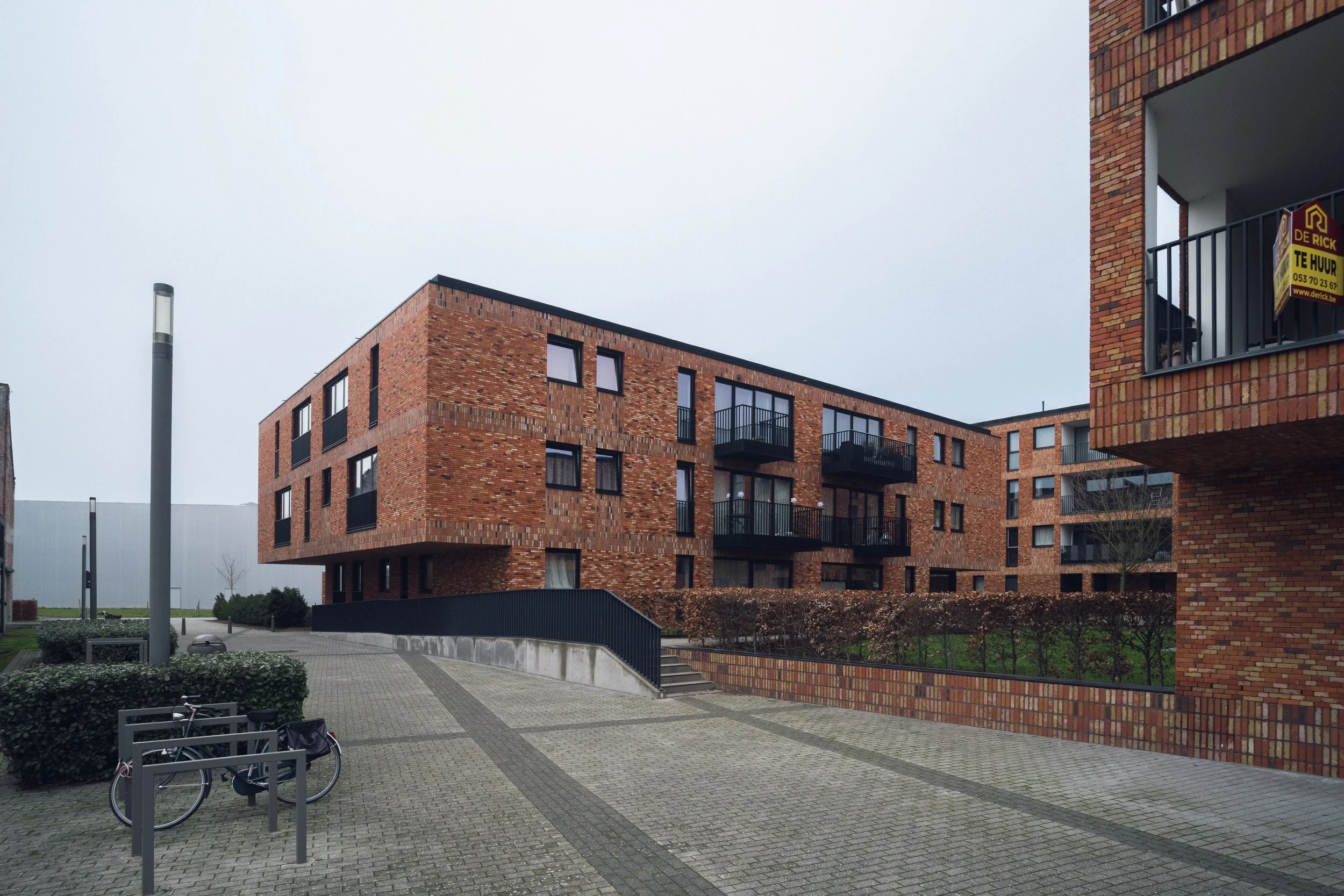 Modern brick apartment complex with balconies and a paved courtyard, bike rack, and street lamps in foreground.