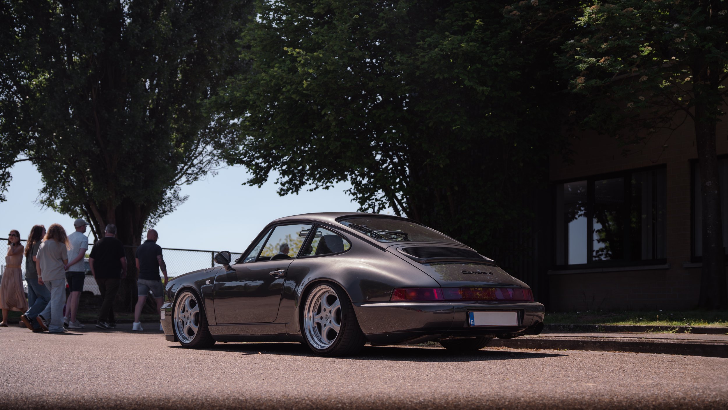 A black vintage Porsche 911 Carrera 4 parked on a street with a group of people and trees in the background.