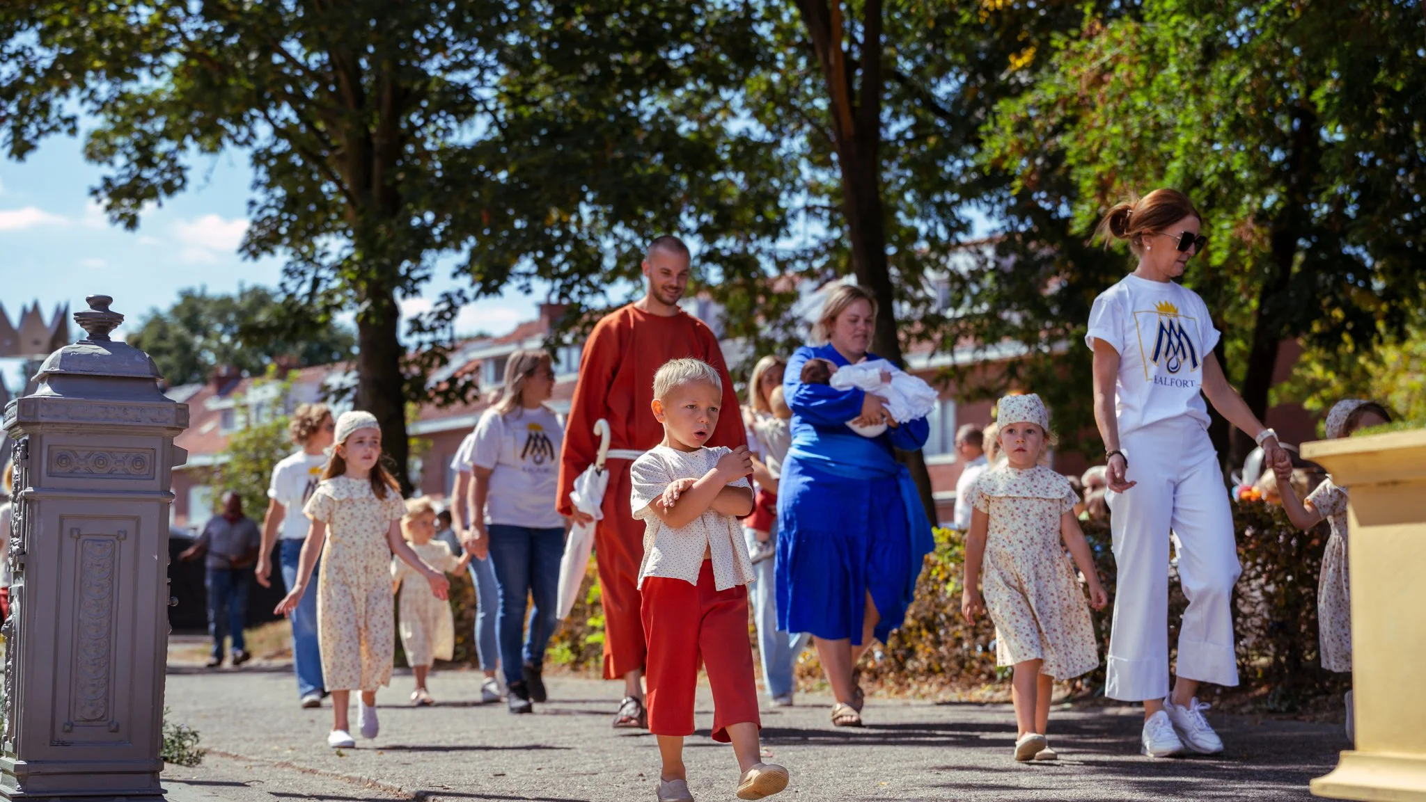 Group of children and adults walking outside on a sunny day, some holding hands, in a suburban neighborhood with trees and houses in the background.