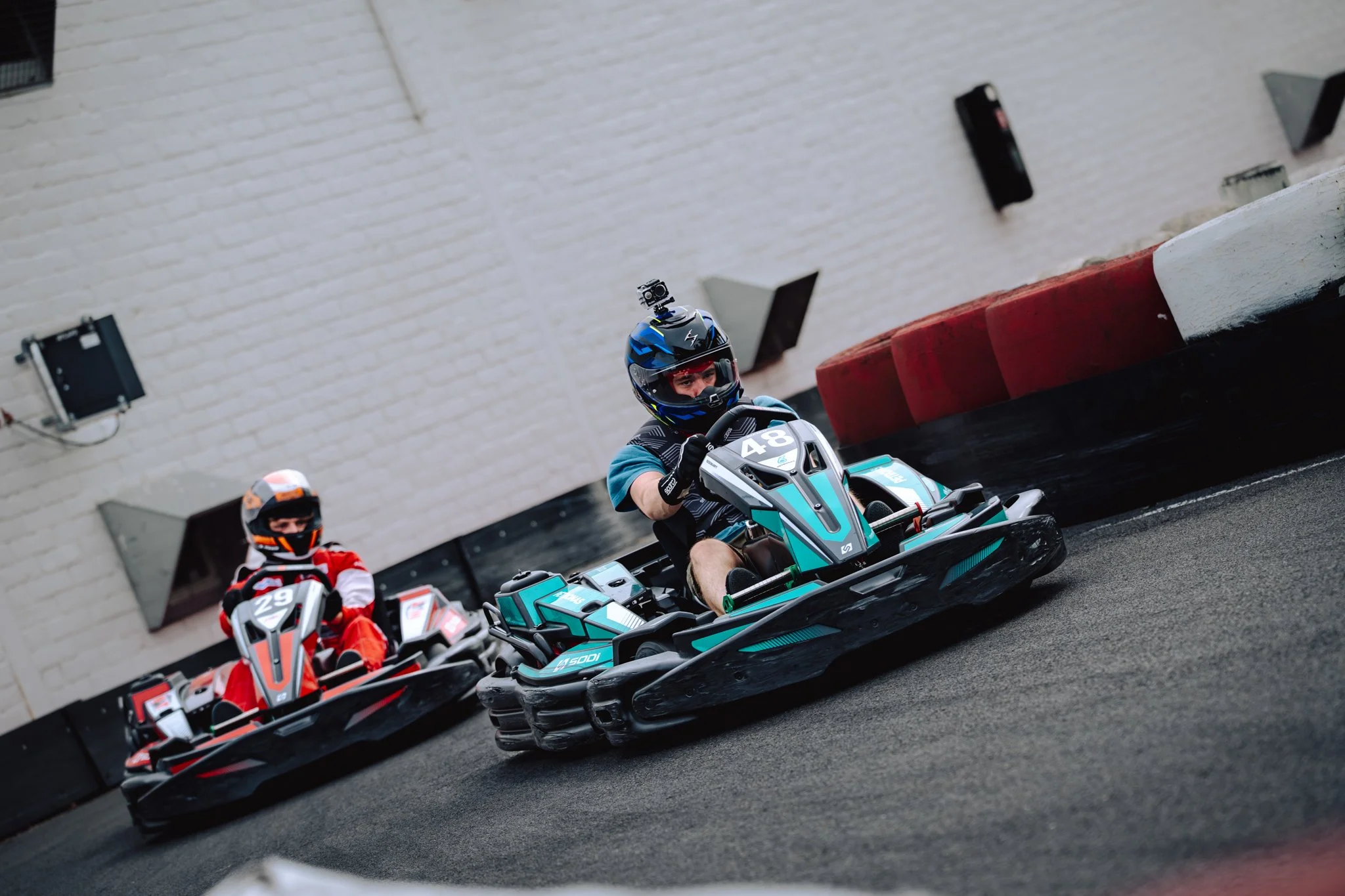 Two people racing on go-karts on an indoor track with white brick walls and safety barriers.