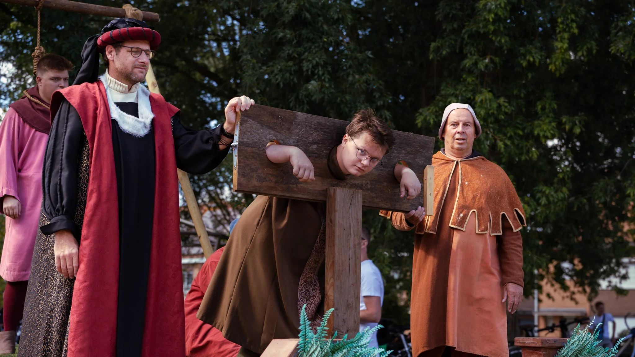 People dressed in medieval costumes participating in a historical reenactment outdoors on a wooden gallows with a person pretending to be executed, surrounded by trees.