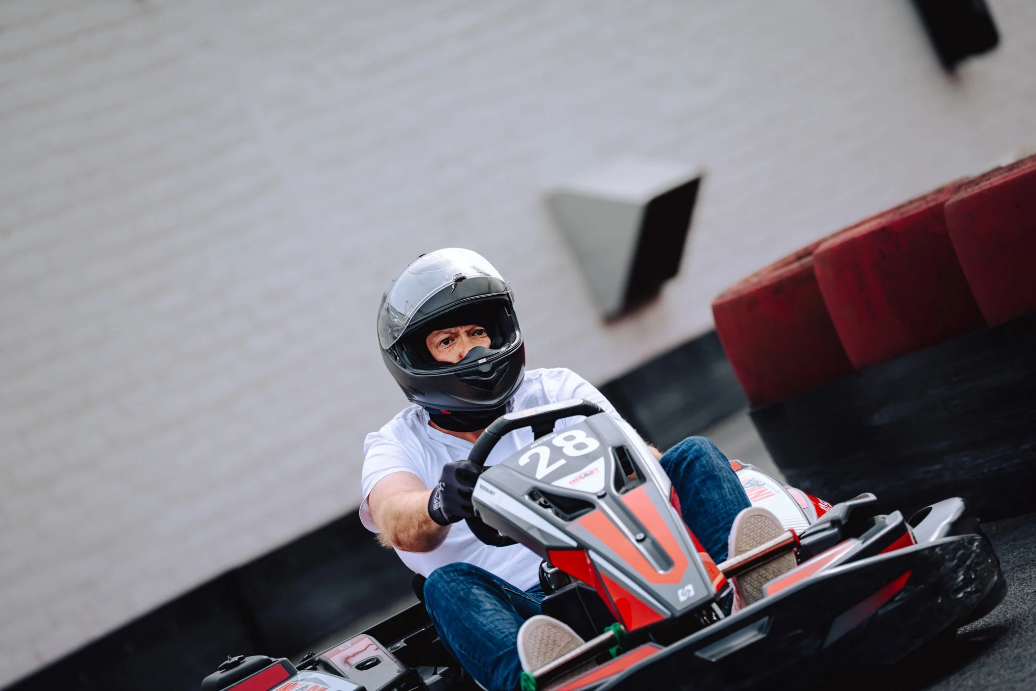 A man wearing a black helmet, white t-shirt, and jeans driving a go-kart with the number 28 on it on a race track.