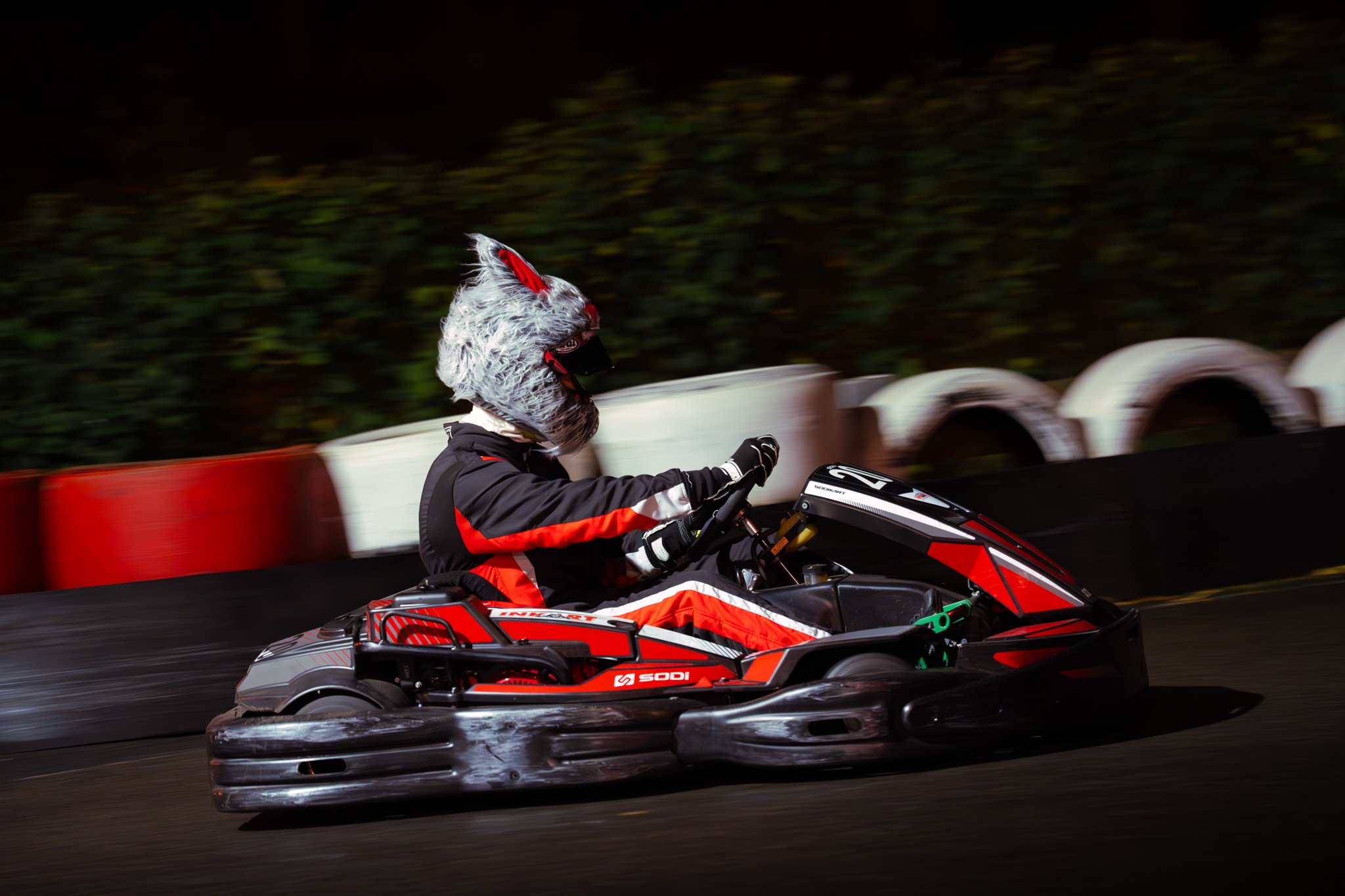 Person racing go-kart on an outdoor track at night, wearing a wolf costume helmet and racing gear