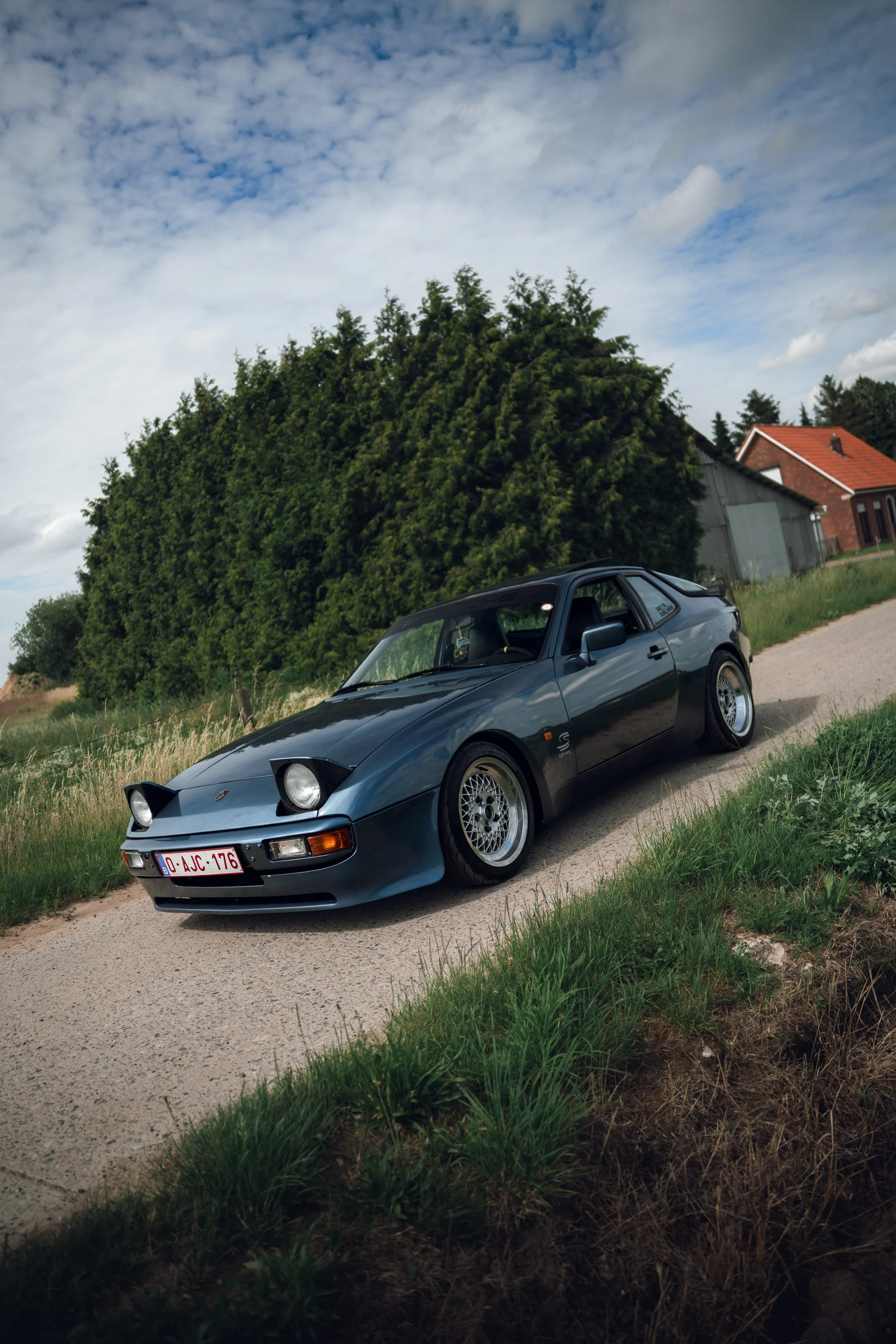 A vintage black sports car parked on a dirt road next to grassy area, with green trees and buildings in the background, under partly cloudy sky.