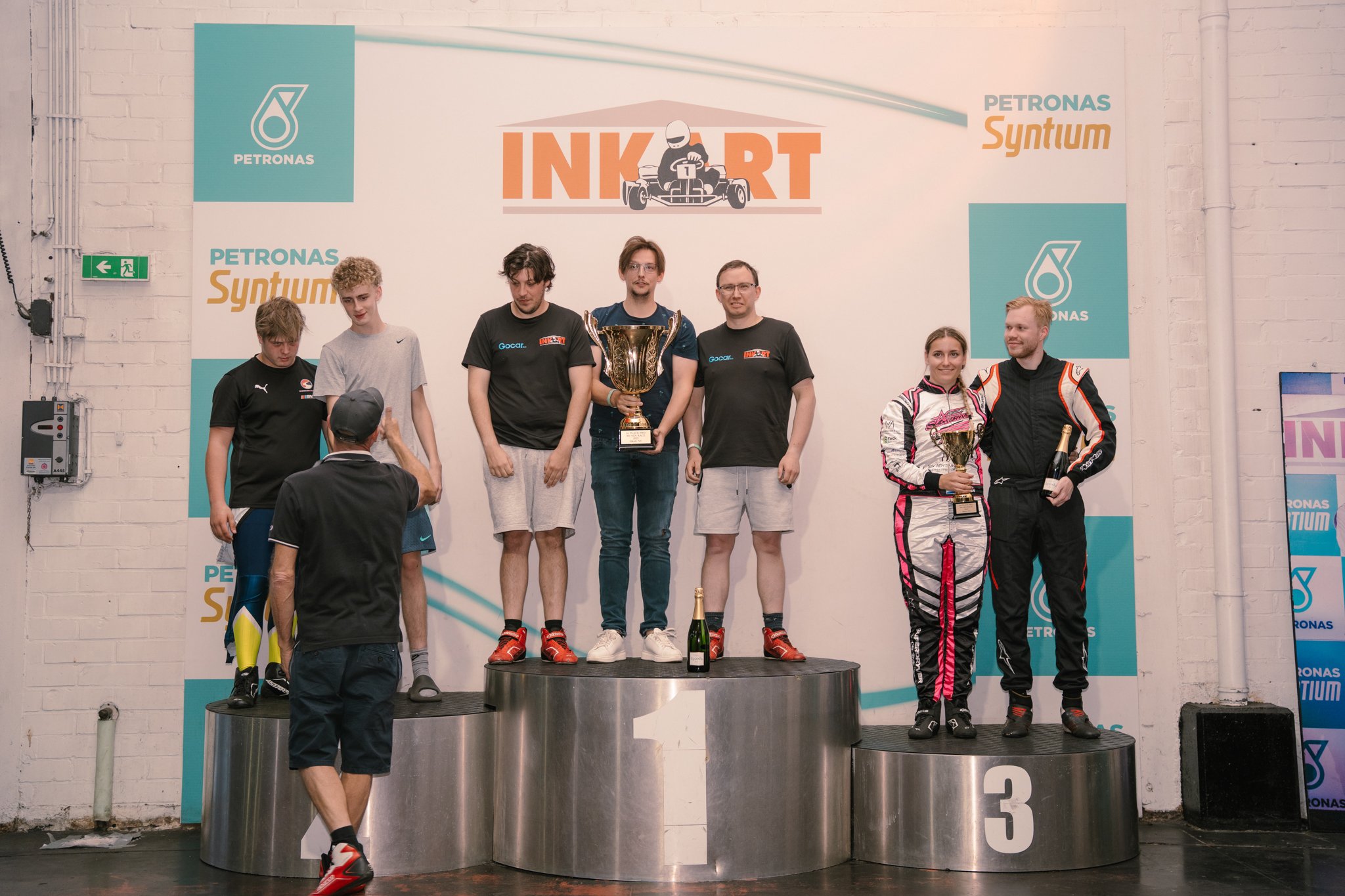 People standing on a winners' podium holding trophies after a go-kart race award ceremony at INKART event, with a backdrop featuring Petronas Syntium branding.