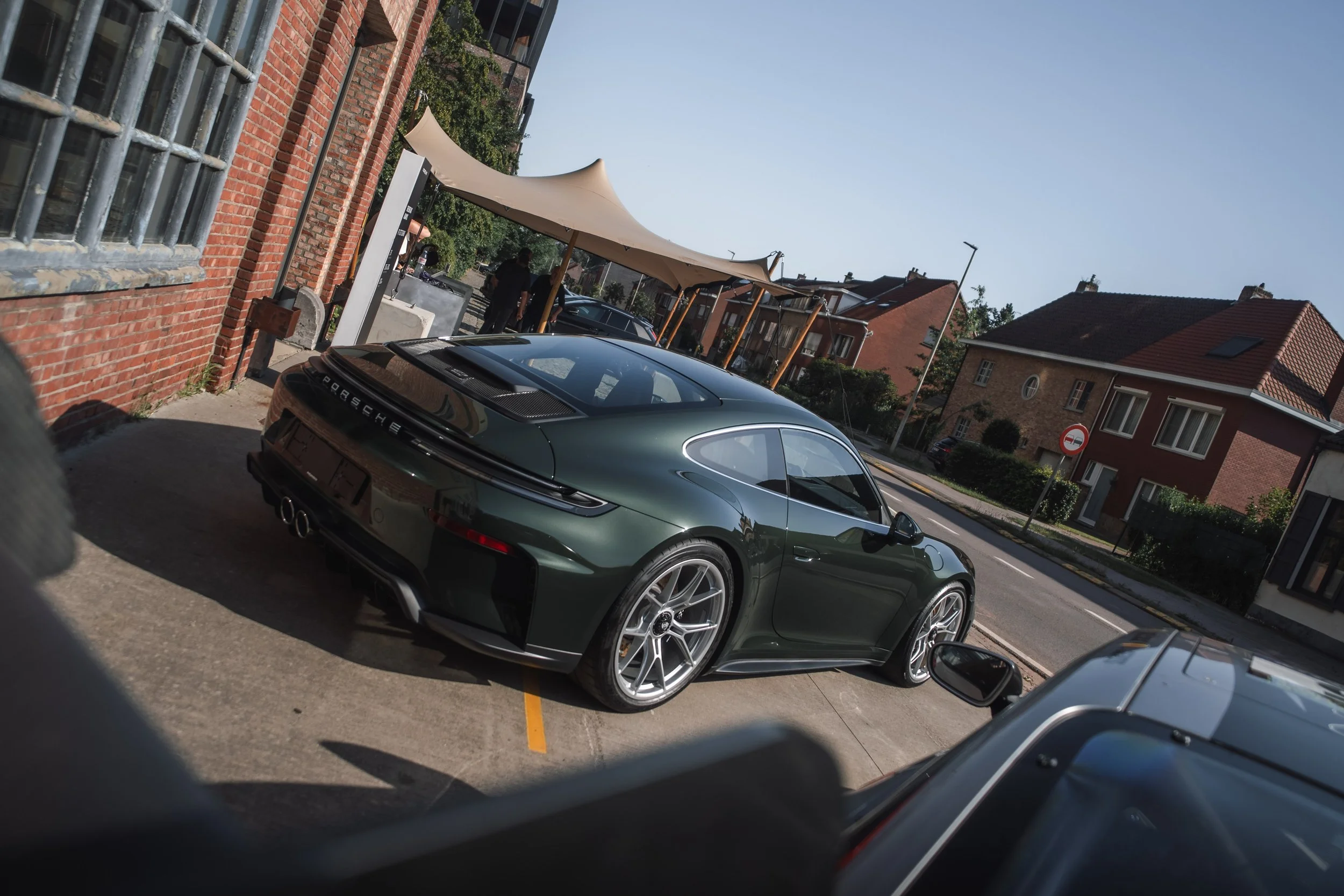 A dark green Porsche sports car parked in an outdoor lot next to a brick building with metal barred windows. There are other cars and people in the background, with a large beige canopy providing shade over part of the parking area. Residential build