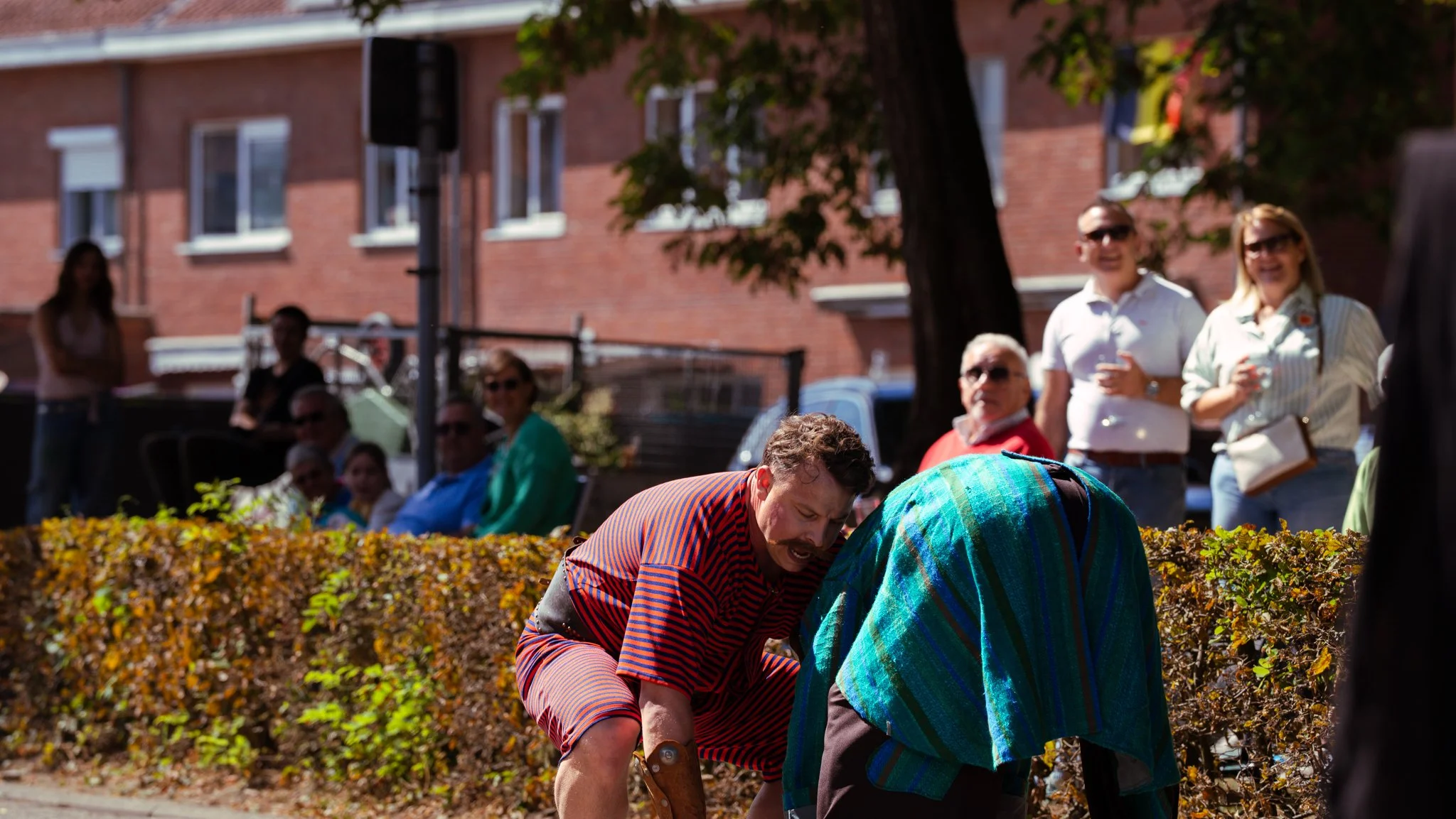 Two men in costume appear to be performing a scene outdoors, with one leaning forward and the other with a hood covering their head, while a group of people watches from a bench and standing in the background.