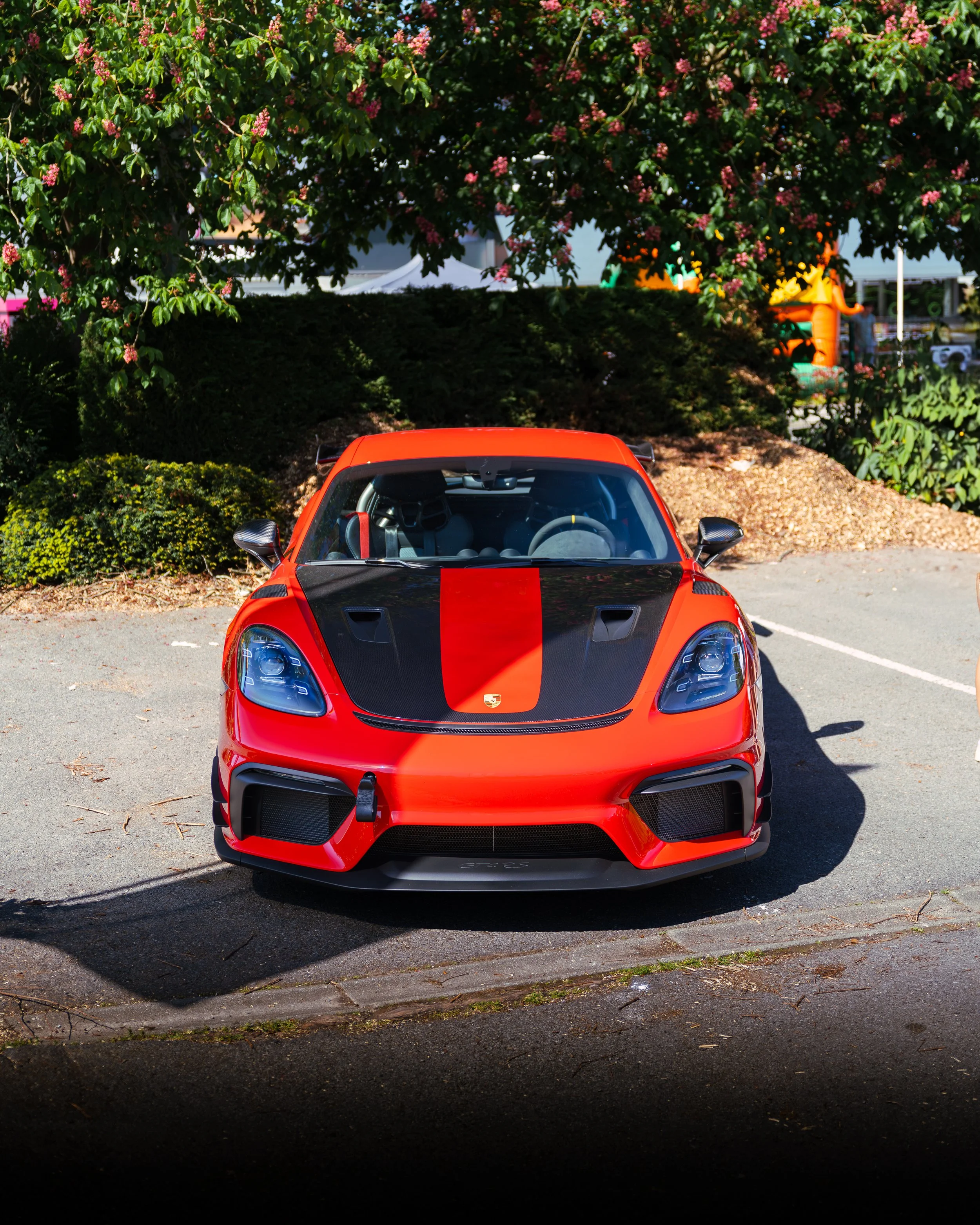 Red sports car with black hood and racing stripe parked in a lot partially shaded by trees.