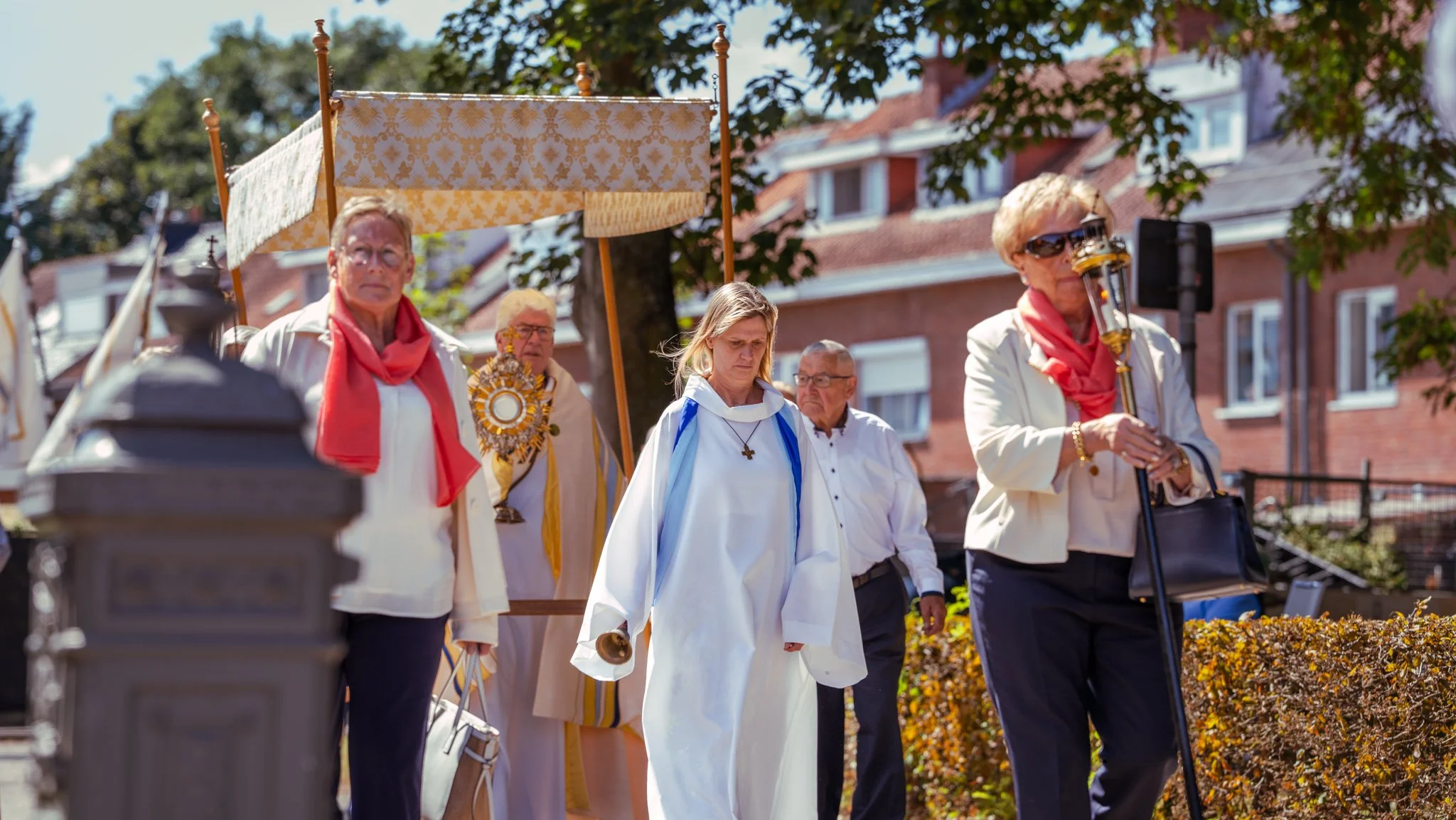 Group of older adults participating in a religious procession outdoors, some dressed in white robes with red scarves, carrying religious symbols, and walking along a sidewalk lined with bushes and buildings in the background.