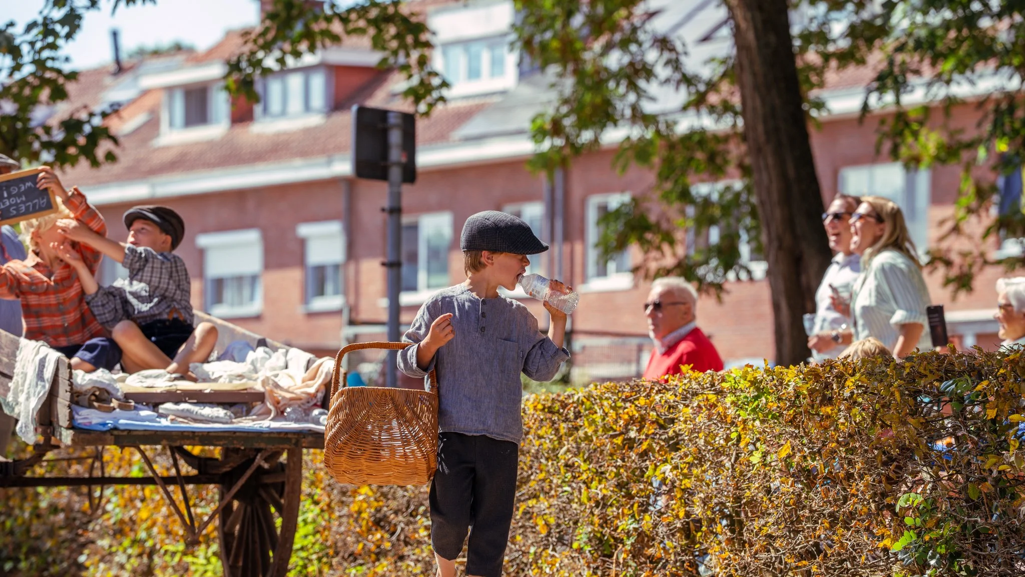 Children and adults outdoors during a sunny day, with children sitting on a cart, a young boy drinking from a water bottle, and adults chatting behind a hedge.