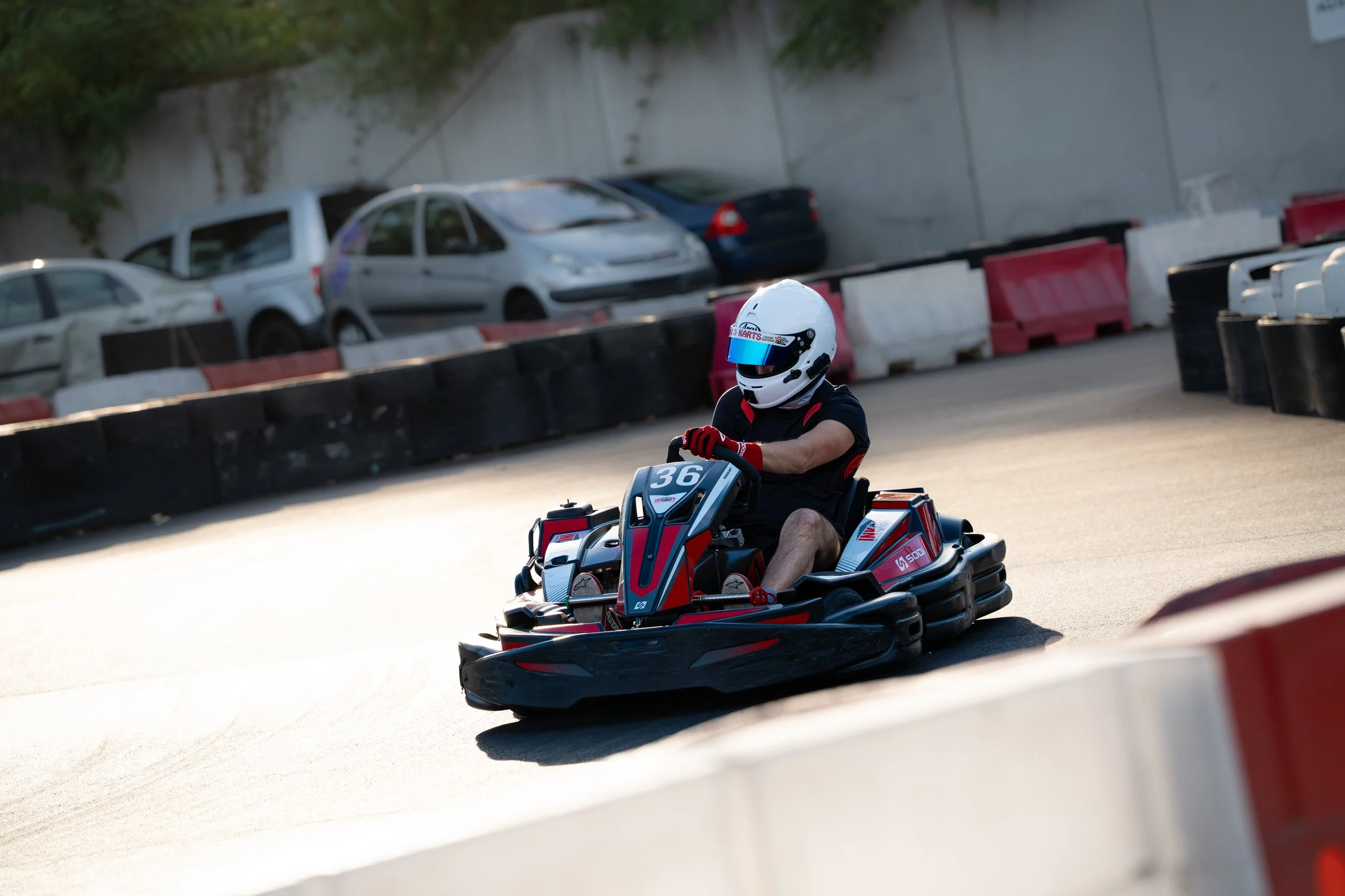 A person wearing a white helmet and black shirt riding a go-kart on a race track, with cars parked behind a barrier and greenery in the background.
