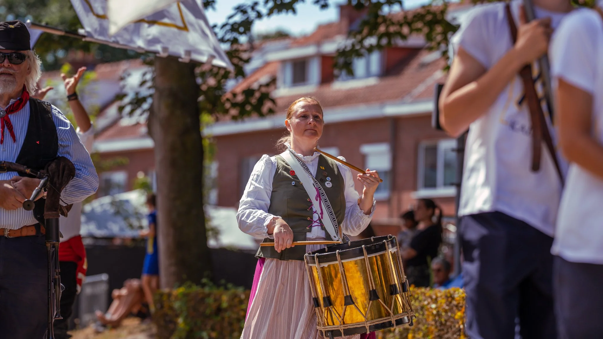 A woman dressed in traditional folk attire playing a drum at an outdoor event, with other people and a brick building in the background.