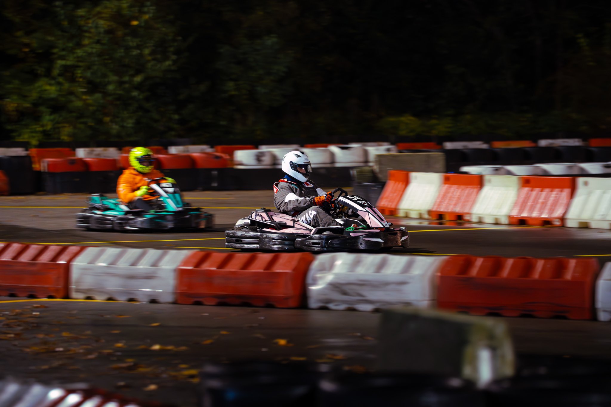 Two people racing go-karts on an outdoor track at night, with barriers and trees in the background.