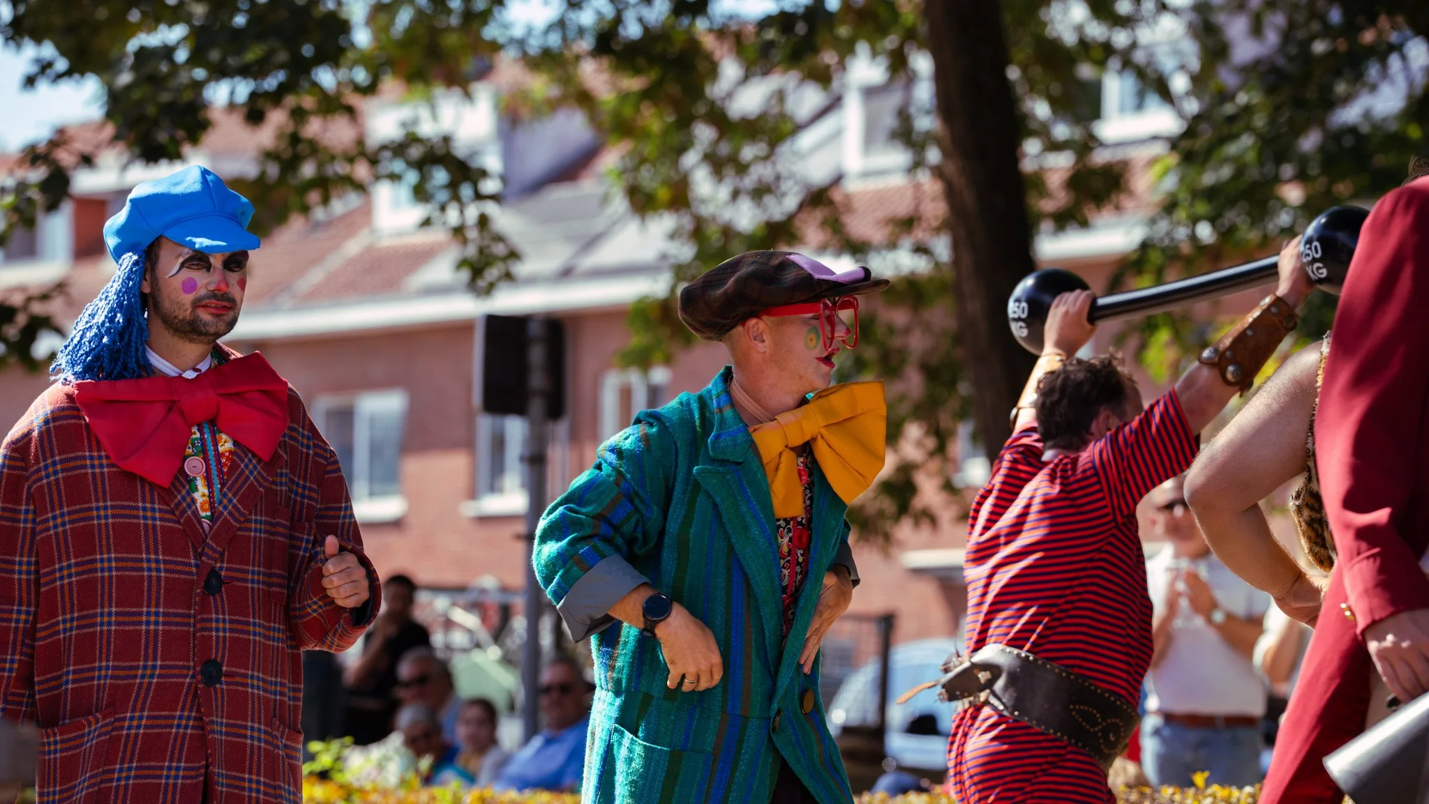People dressed as clowns at an outdoor event, some holding objects, with a background of trees and buildings.