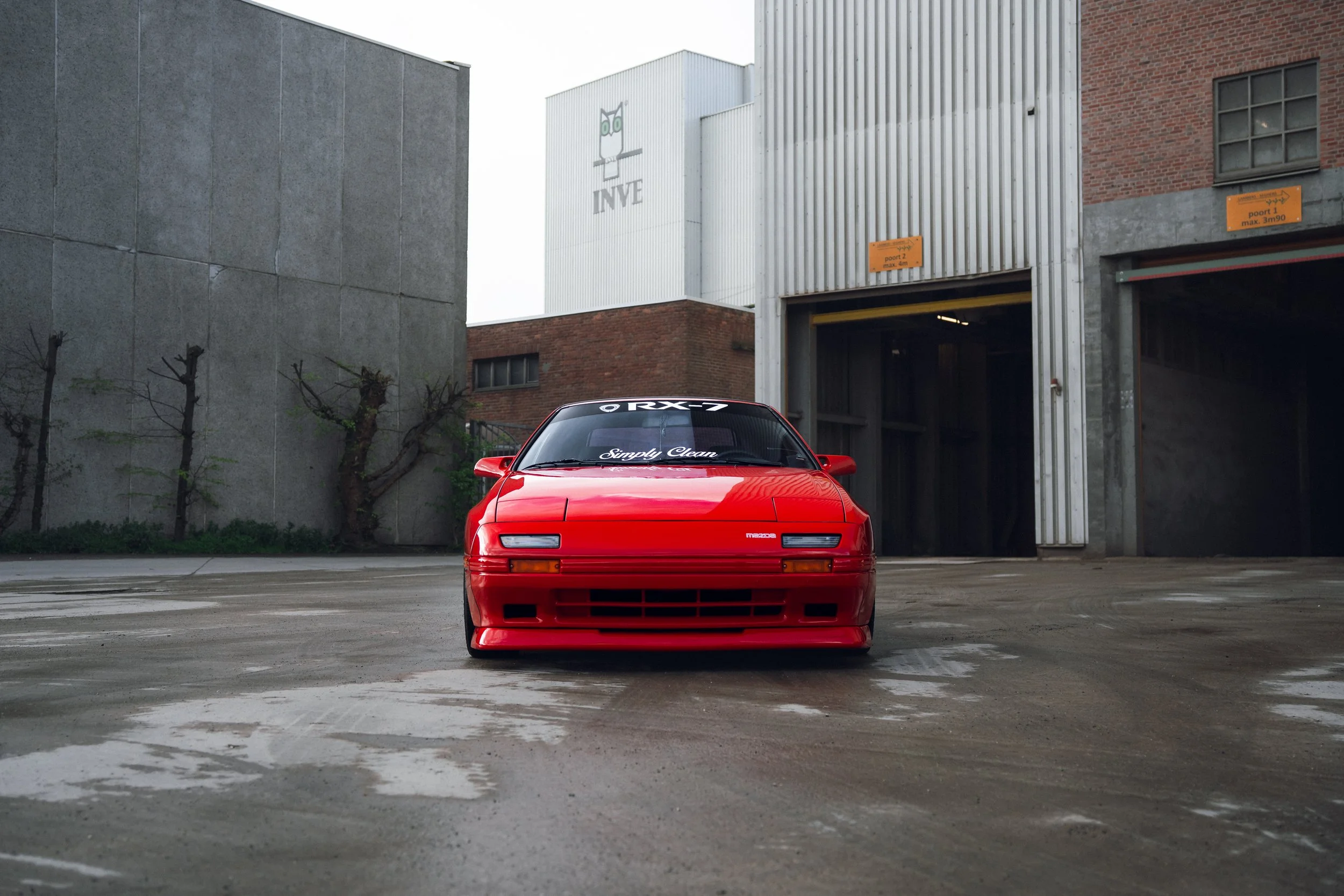 Red vintage Mazda RX-7 sports car parked in an industrial area with concrete and metal buildings.