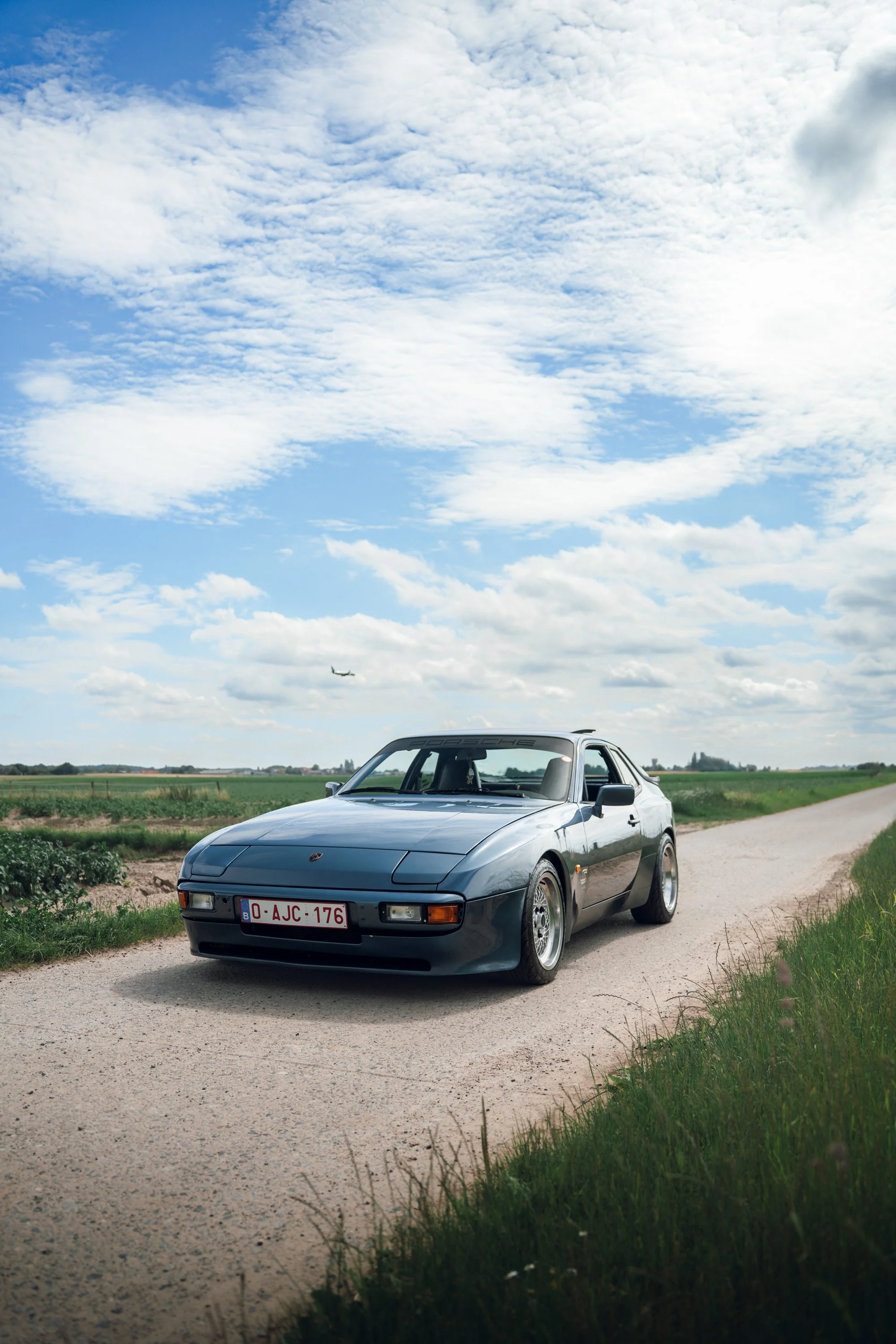 A silver Porsche sports car parked on a gravel road in a rural area with green fields and a cloudy sky. A plane is visible flying in the background.