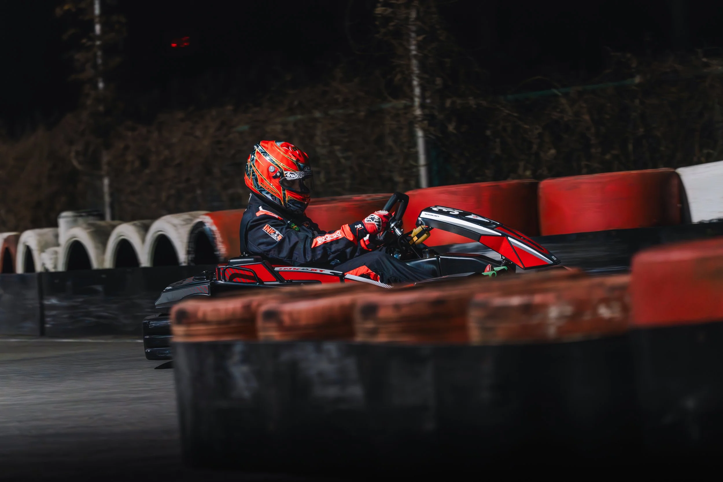 Nighttime go-kart race featuring a racer in a red and black helmet and racing suit, navigating a track with red and white barriers.