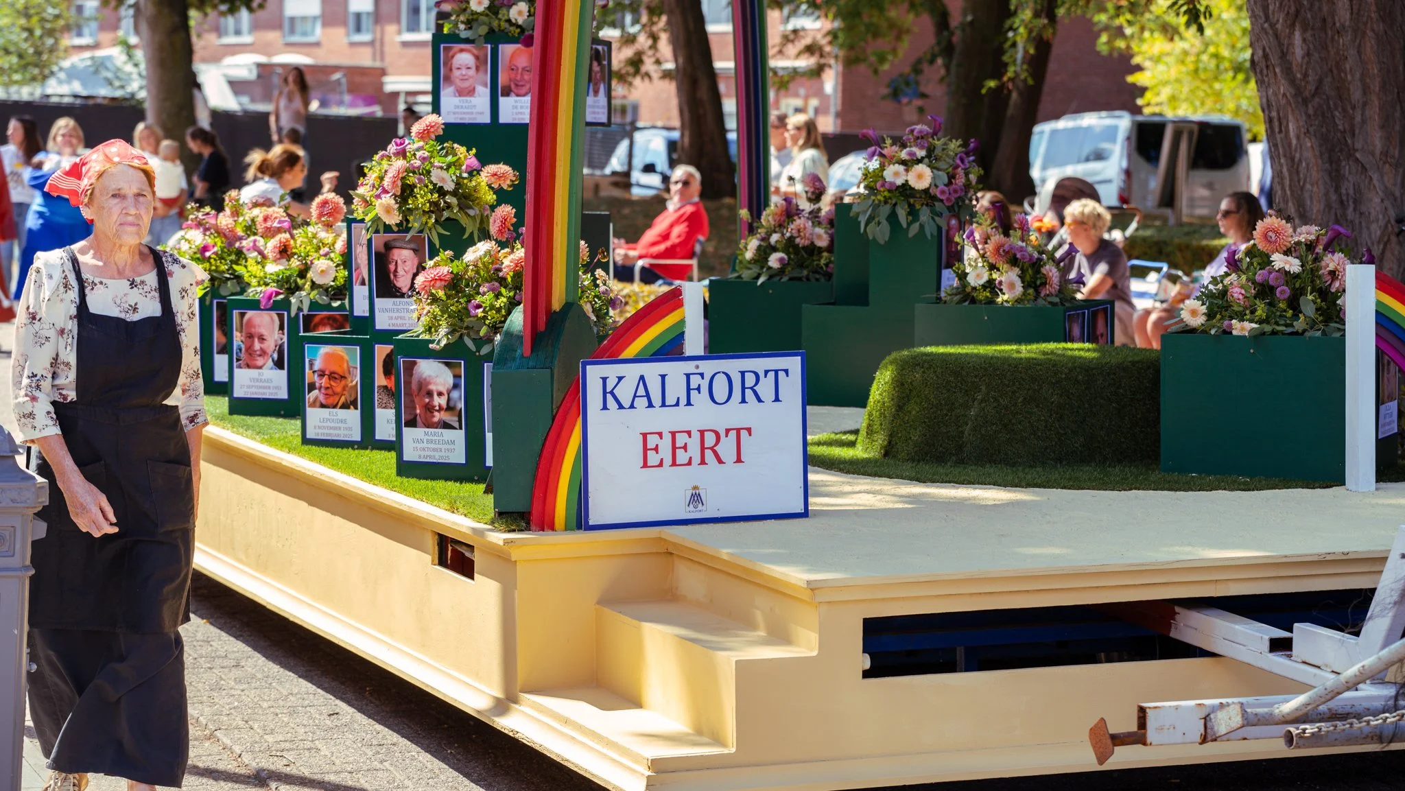 A float decorated with flowers and photos of people, with a sign reading 'Kalfort Eert' and a rainbow decoration. There are several people sitting and standing in the background.