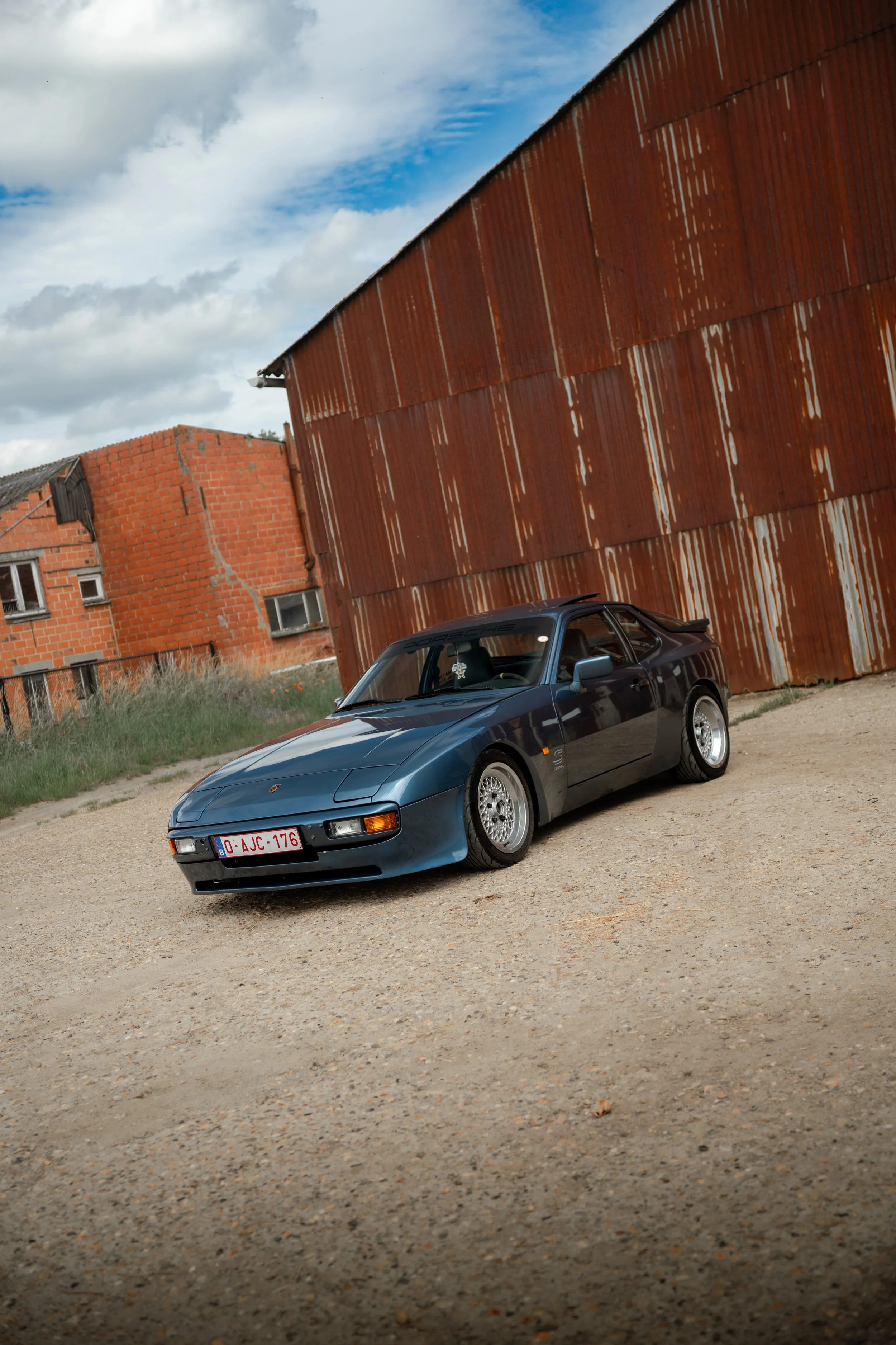 A black sports car parked on a dirt ground next to an old rusty building with a corrugated metal exterior. There are other buildings in the background under a partly cloudy sky.